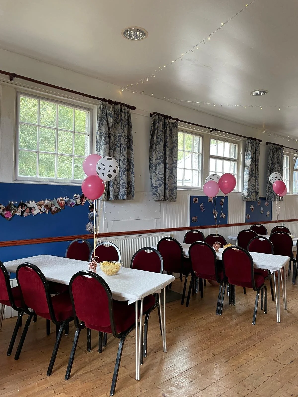 Decorated indoor dining area with tables, red chairs, pink and black-and-white balloons, photo garland, and string lights, likely set up for a celebration.