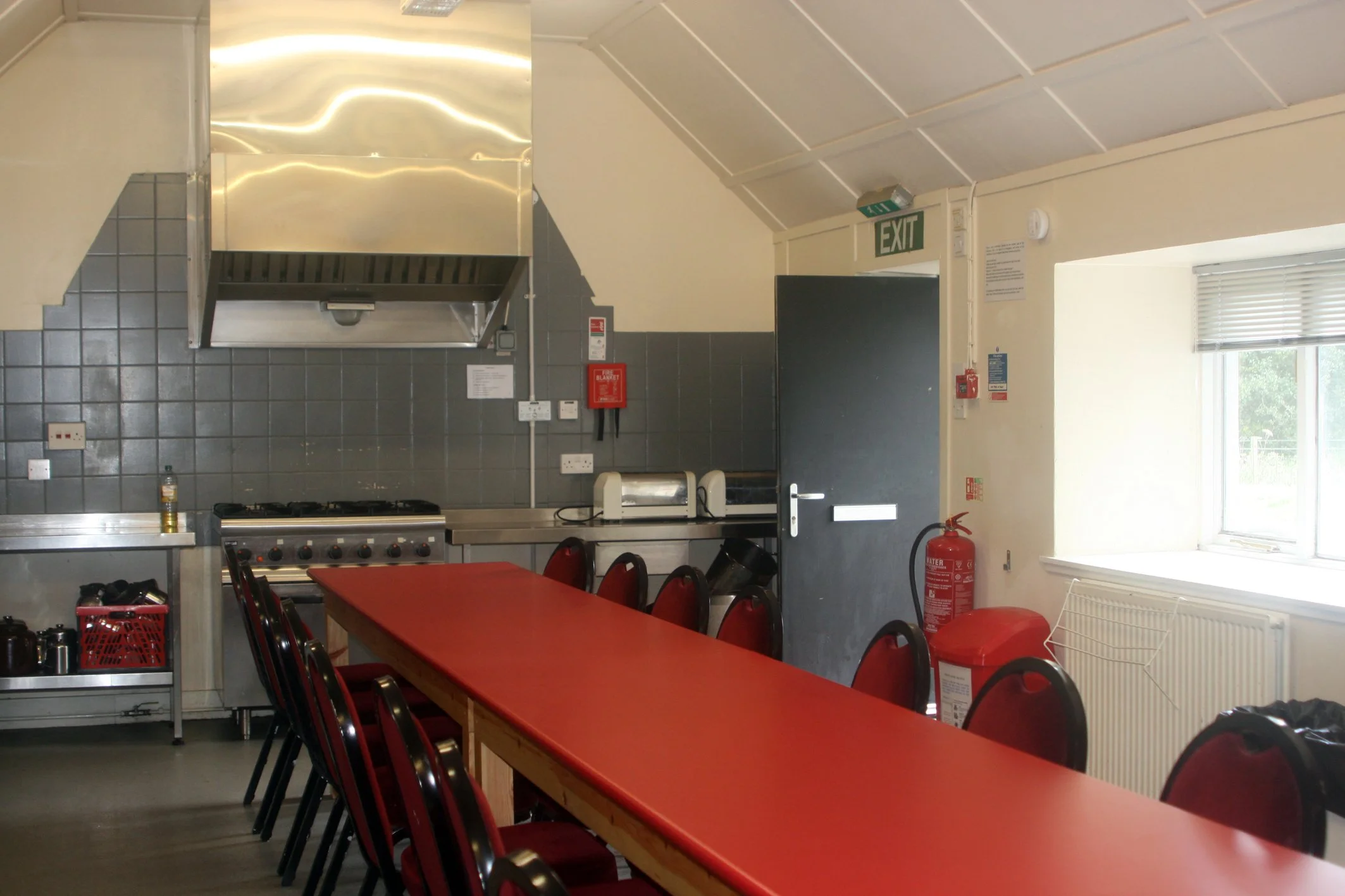 A view of a kitchen with a red table, black chairs, and kitchen appliances including a stove with a stainless steel hood, a toaster, and a microwave. The kitchen has gray tiled walls, white walls, a window with blinds, and an exit door with an illuminated exit sign above it. There is a red fire extinguisher and a blue sticker near the door.