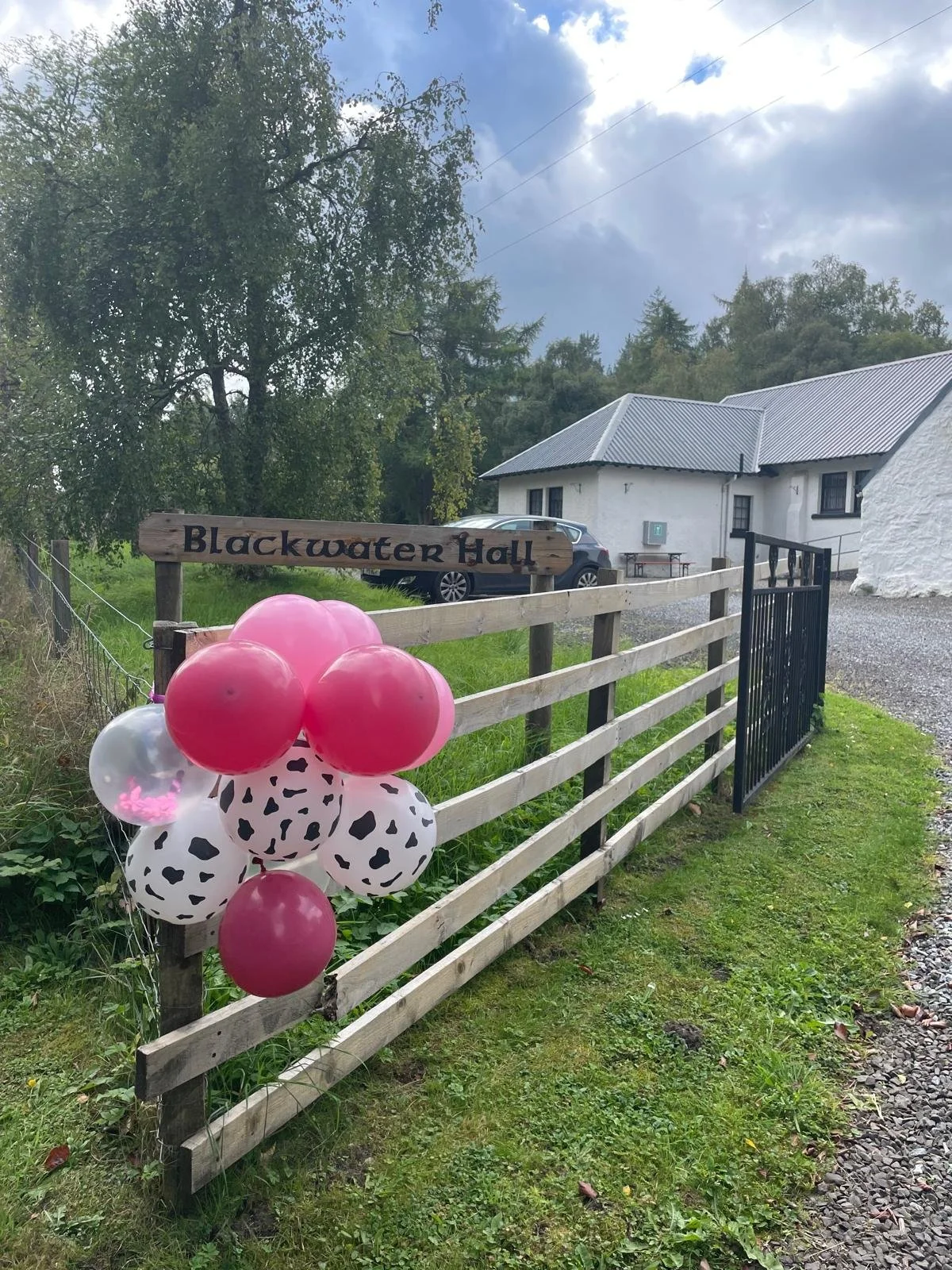 A wooden fence with a sign that reads "Blackwater Hall." Decorated with pink and black-and-white spotted balloons, alongside a white hall with a roof and car park, set in a rural area with trees and cloudy sky in the background.