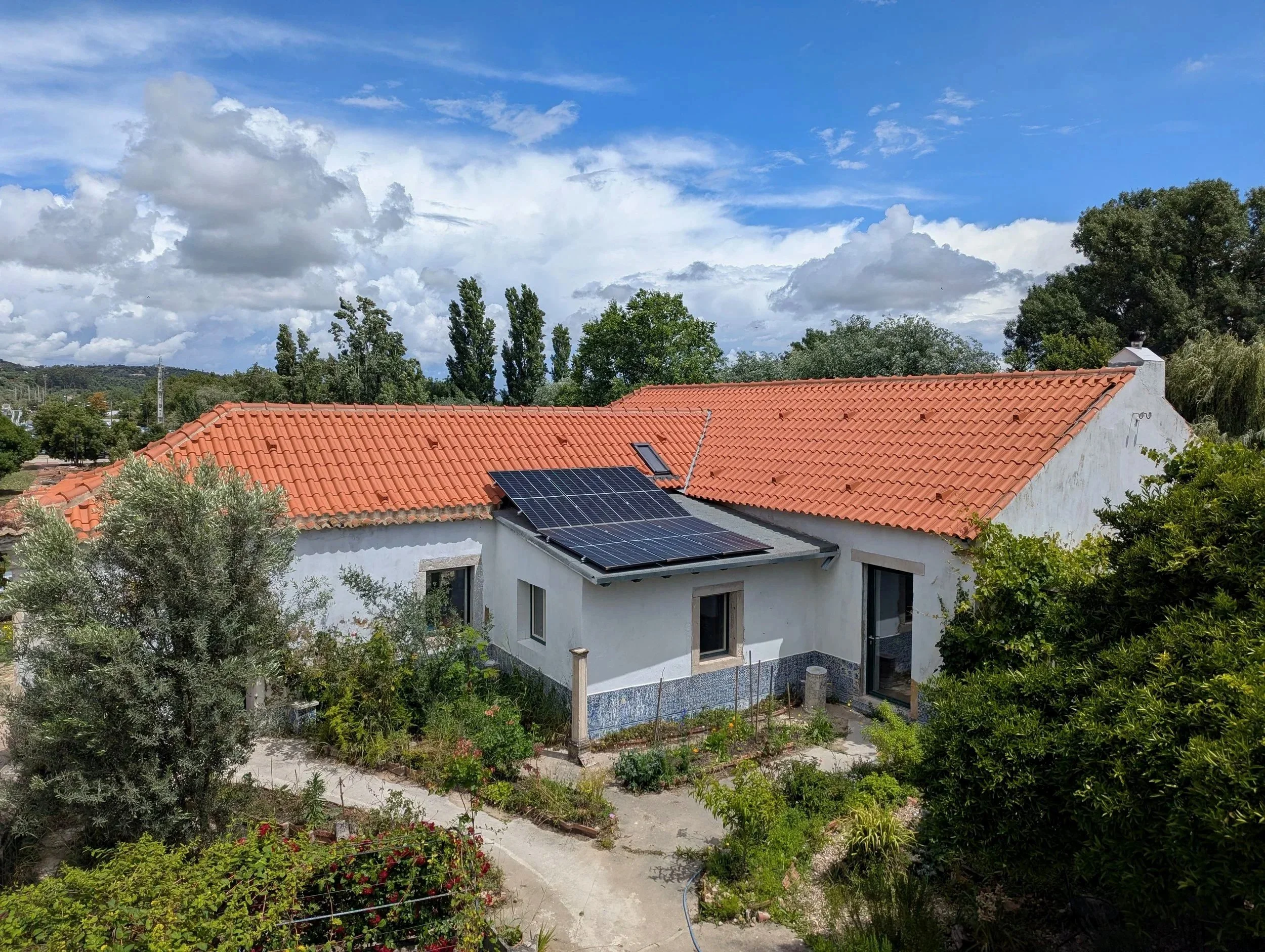A house with a red tiled roof and solar panels, surrounded by trees and a garden, under a partly cloudy sky.