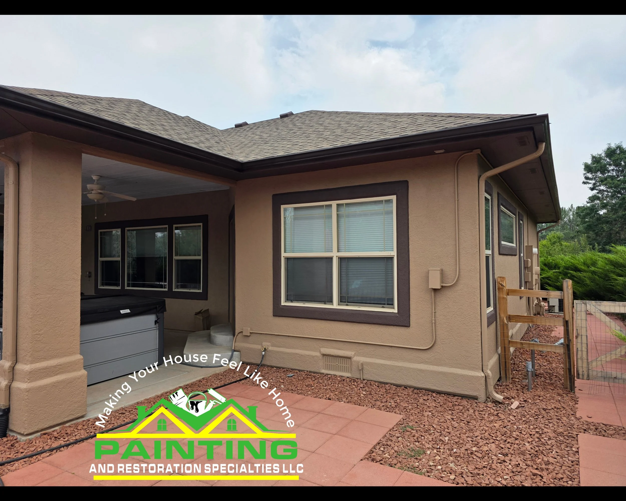 Backyard patio area of a house with window, ceiling fan, and covered porch. There is a garage door with a garden hose attached and a partially built wooden fence, with trees and cloudy sky in the background.