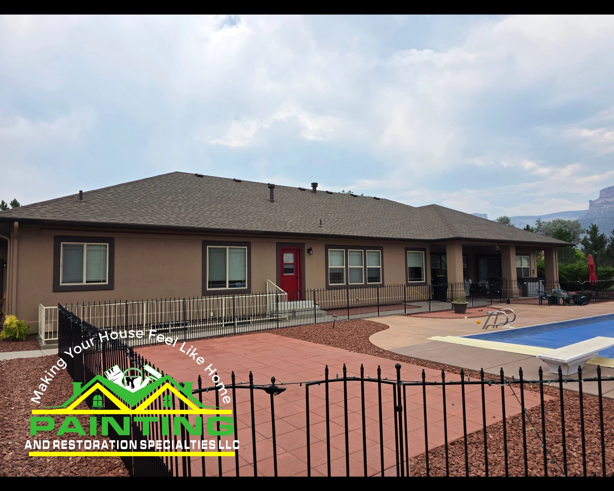 A backyard with a concrete patio, a swimming pool, and a fenced yard. There is a house with beige walls and dark brown trim, and a red door. A sunny sky with some clouds and distant mountains are visible in the background.