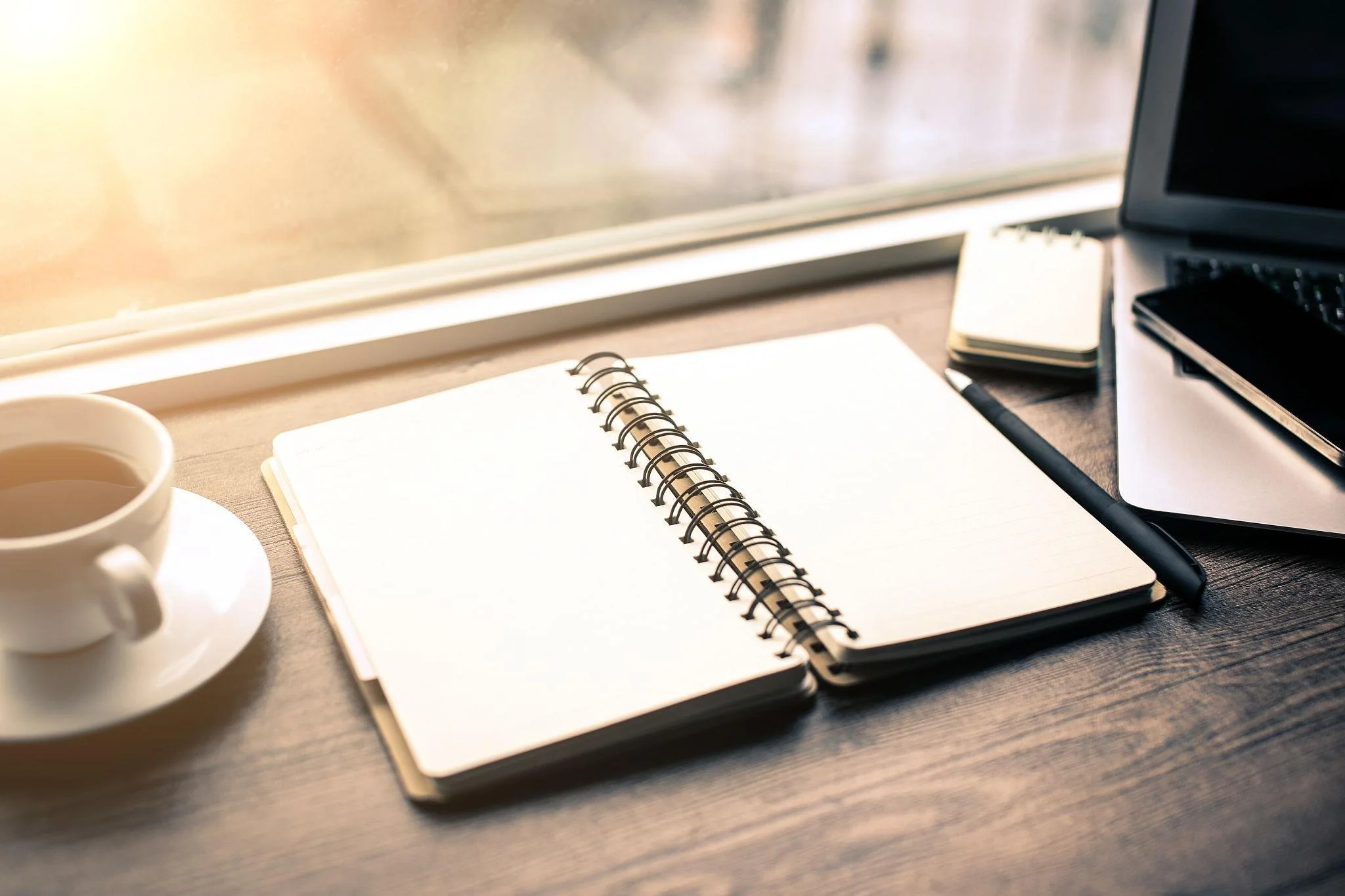 A desk workspace with a cup of coffee, an open spiral notebook, a pen, a laptop, and a stack of sticky notes near a window.