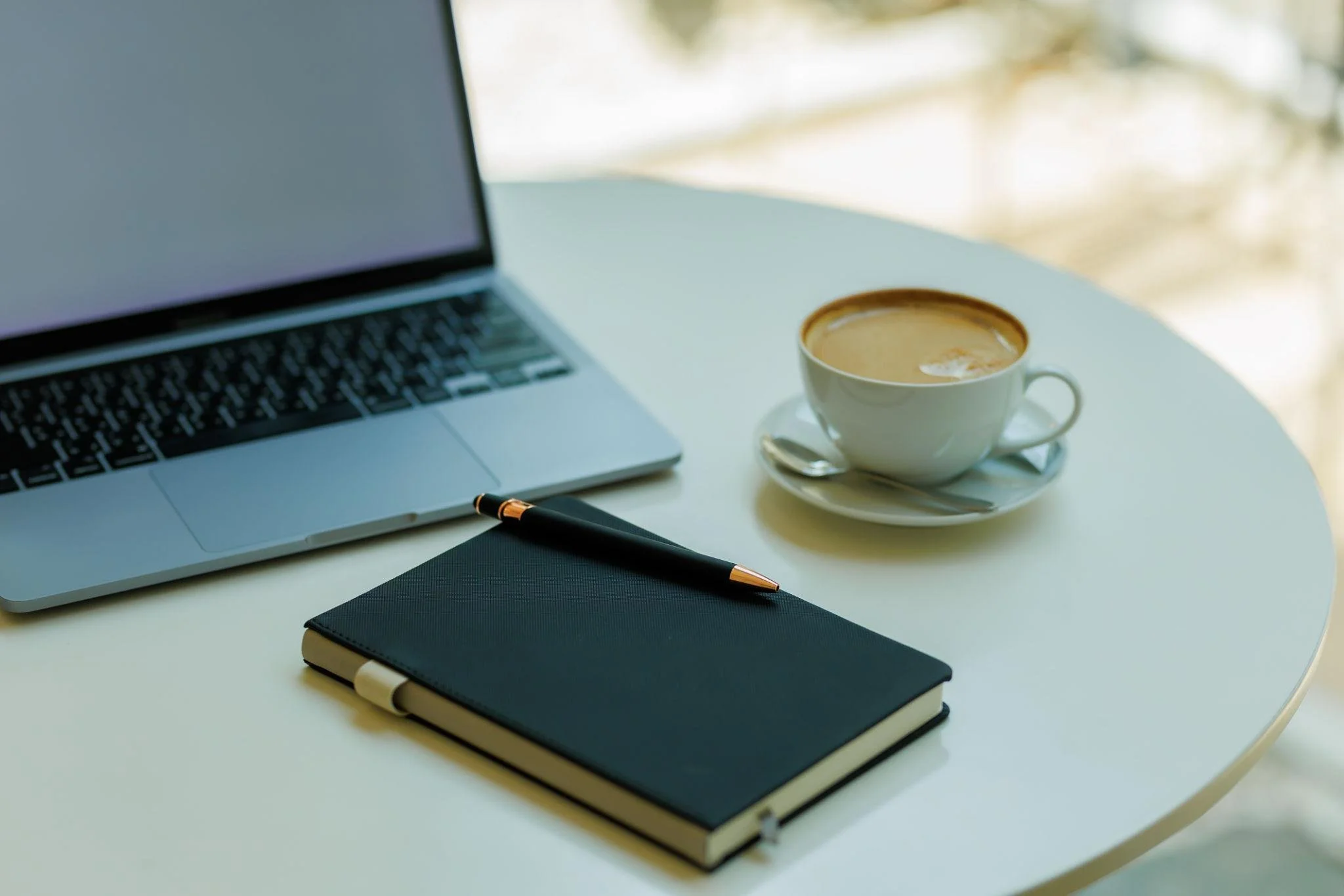A white table with an open laptop, a closed black notebook with a pen on top, a cup of coffee with a spoon on a saucer, and a black mouse pad. A black pen is also visible on the table. The background shows blurred natural light and trees.
