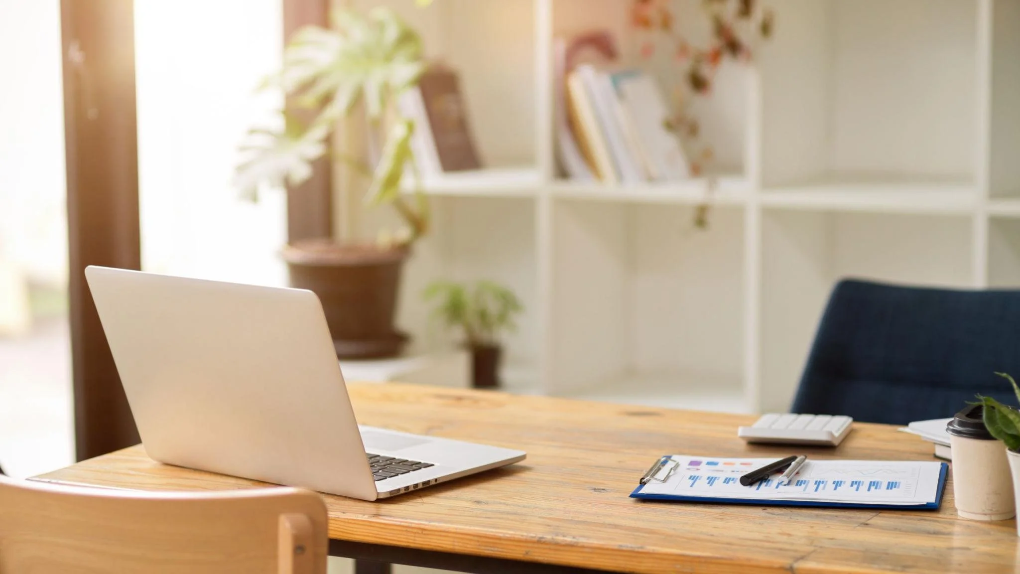 A wooden desk with a silver laptop, a clipboard with a chart, a pen, and a disposable coffee cup in a bright room with shelves, books, and plants.