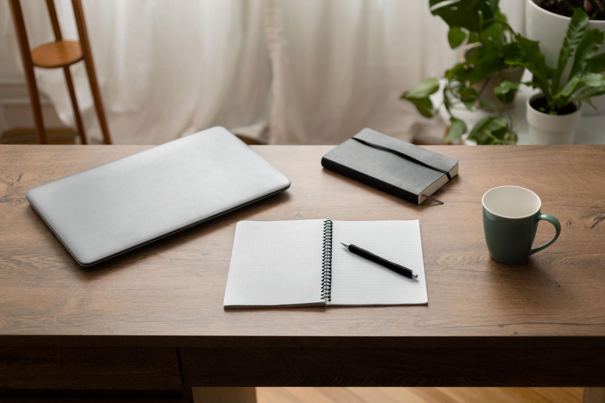 A wooden desk with a closed silver laptop, an open spiral notebook with a pen, a black and white striped notebook, a white coffee mug, and a green potted plant in the background.