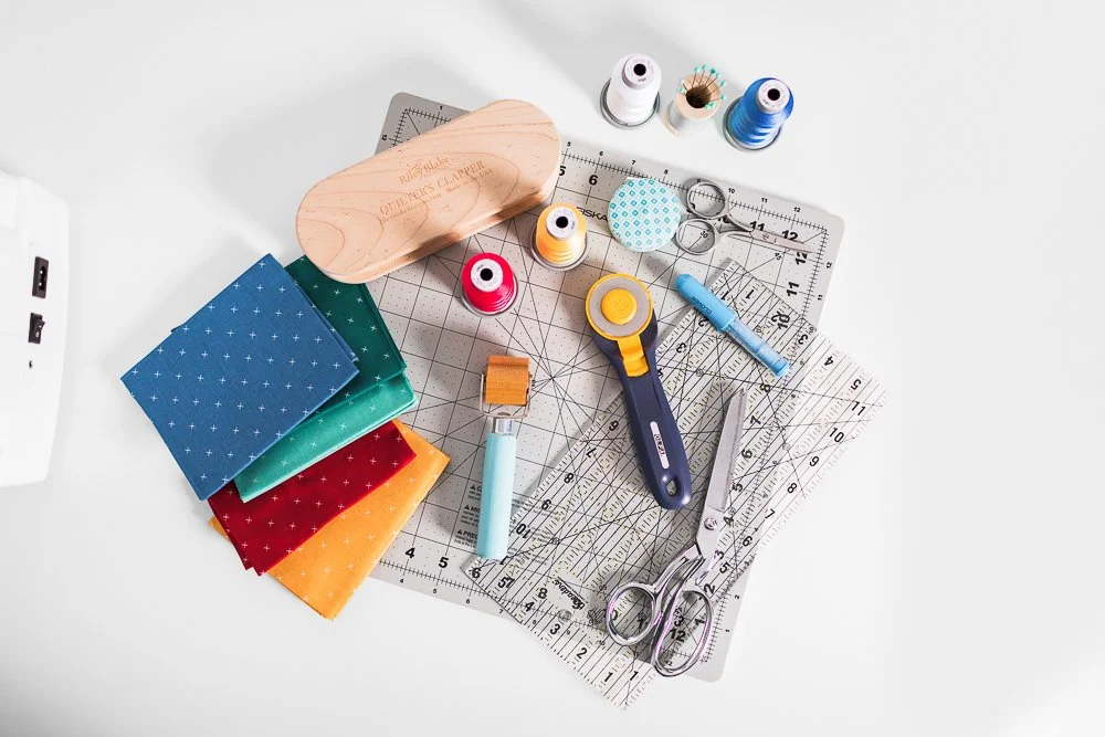 Sewing and fabric tools on a white surface, including patterned fabric, scissors, rulers, pins, thread spools, a rotary cutter, and a cutting mat.