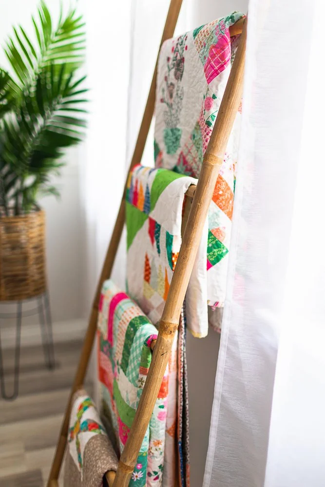 Colorful quilts hanging on a wooden ladder, with a potted plant in the background.