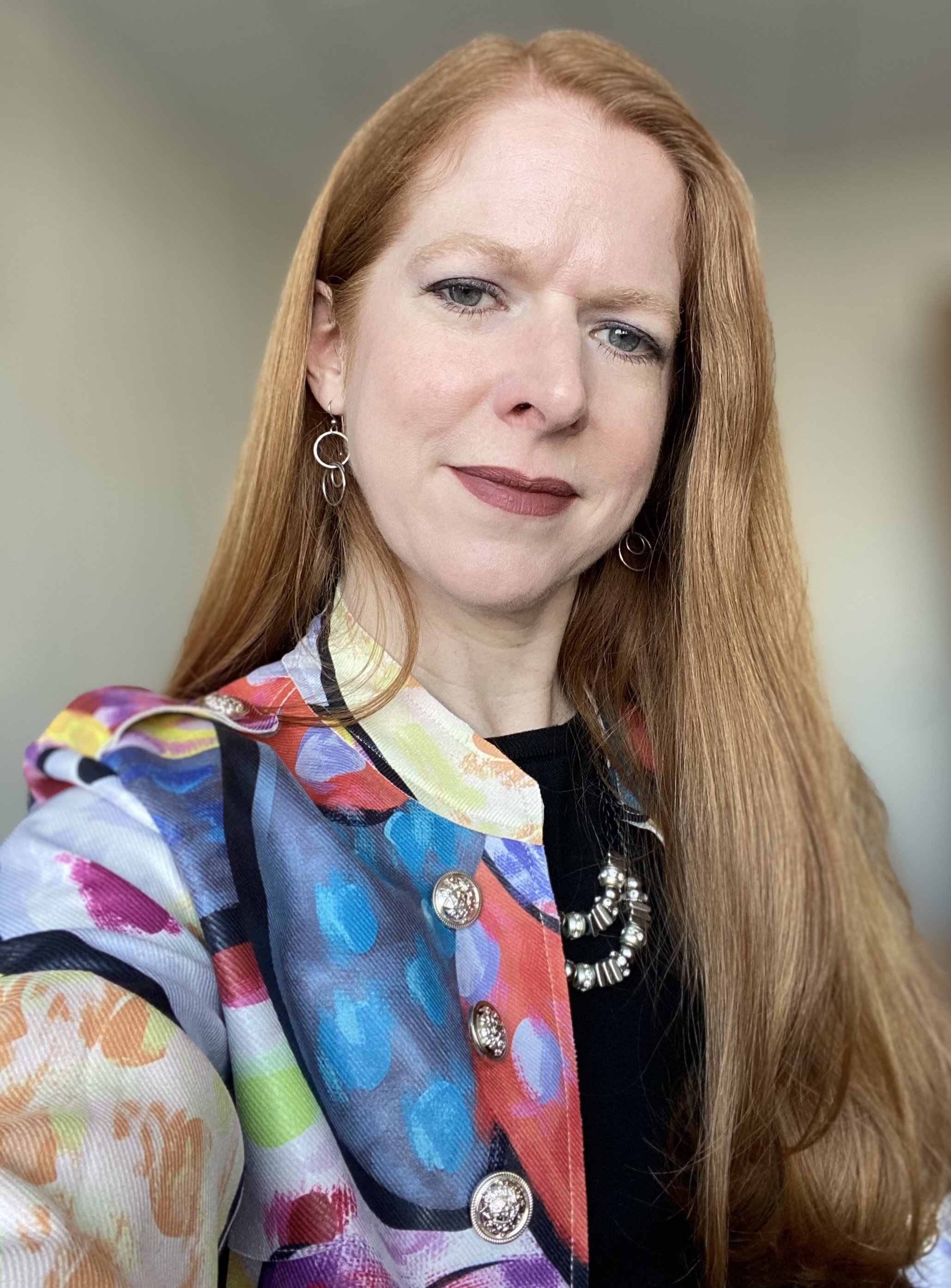 Close-up of a woman with long red hair, wearing colorful clothing, earrings, and a necklace, against a plain background.