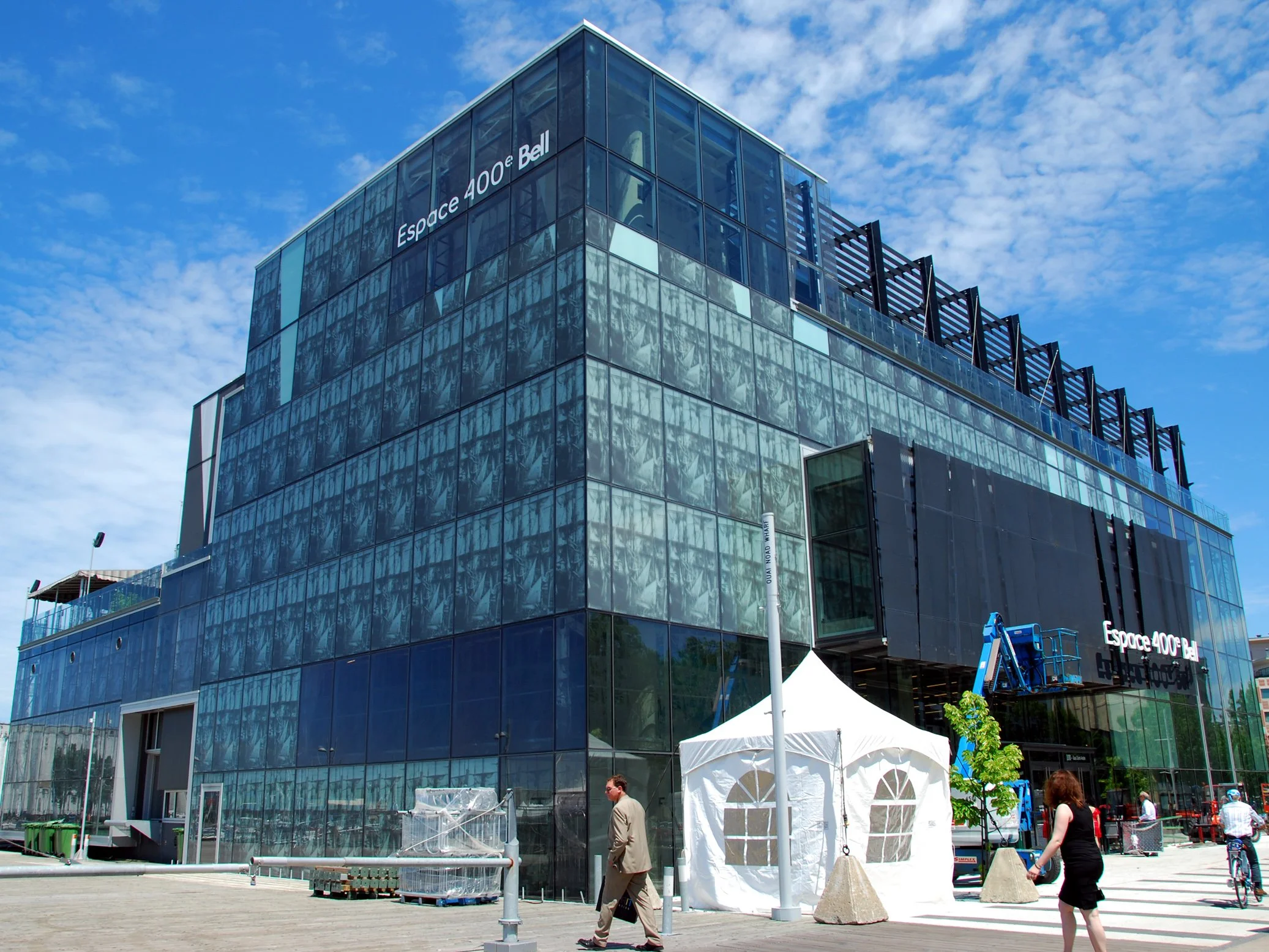 Bâtiment moderne avec façade en verre portant une inscription 'Espace 400e Bell', des gens qui marchent à l'extérieur, une tente blanche et un élévateur pour travaux