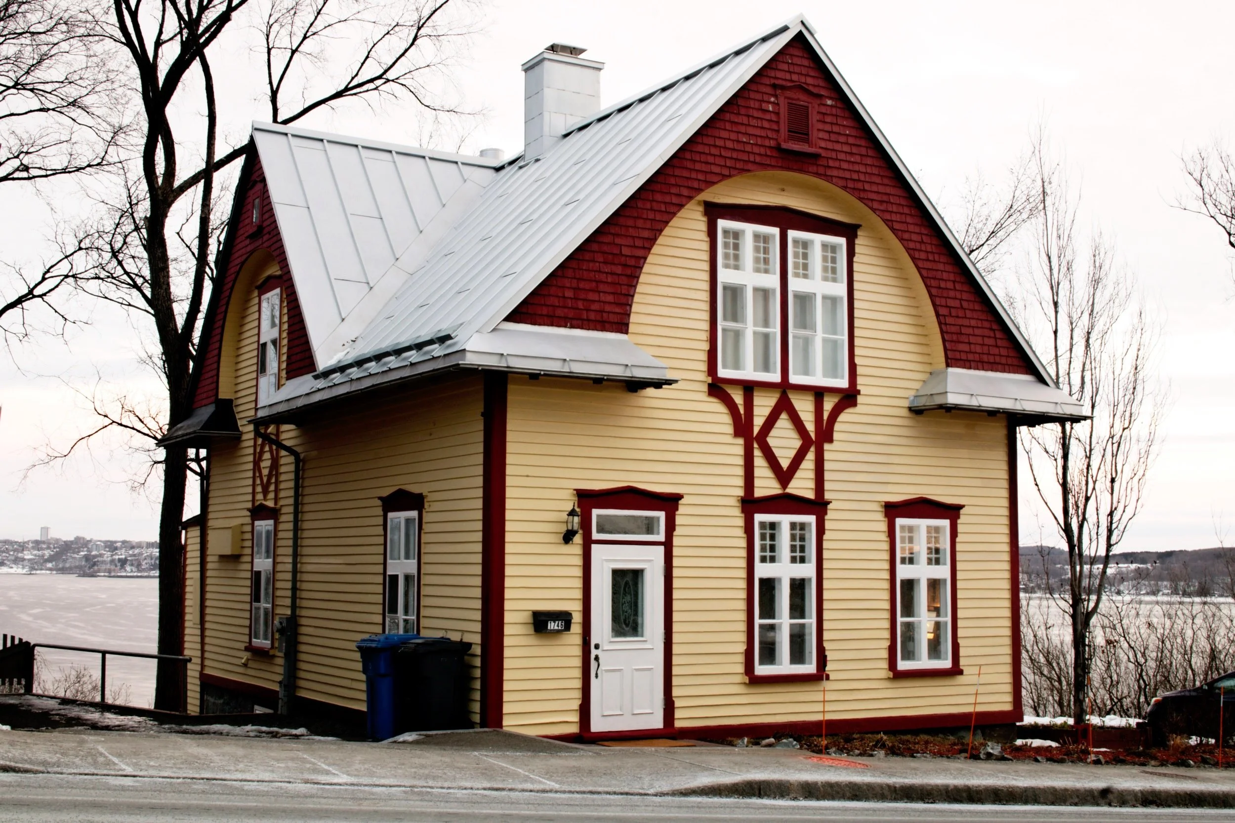 Maison en bois de style scandinave avec façade jaune, détails en rouge, toit en métal et plusieurs fenêtres, située près d'un lac en hiver.