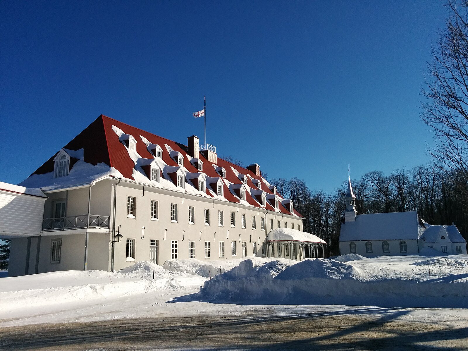 Bâtiment blanc avec toit en pente rouge recouvert de neige, petite église à côté, dans un paysage enneigé avec des arbres nus, ciel bleu clair.