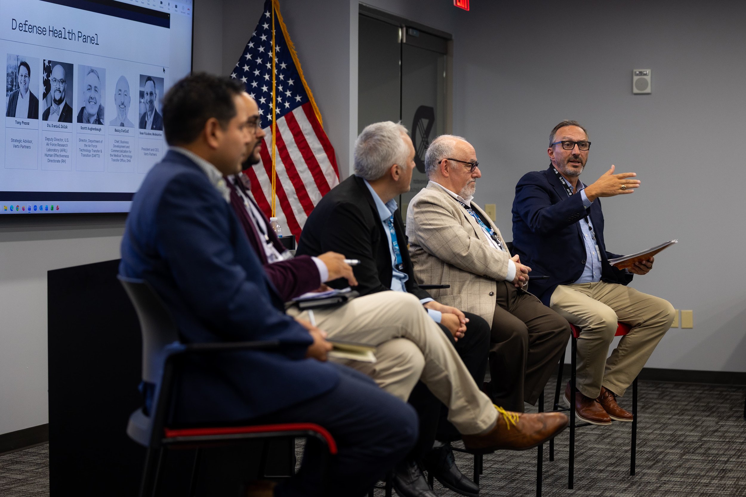 A panel of five men sitting in a row during a conference or meeting, with a presentation slide on the wall behind them and an American flag nearby. One man is speaking and gesturing with his hand.