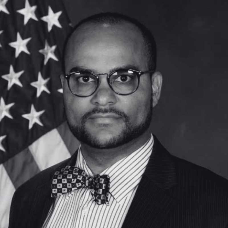 A man with glasses, a suit, and a bow tie standing in front of an American flag.