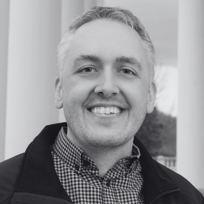 Portrait of a smiling man with short gray hair, wearing a checkered shirt and a dark jacket, standing outdoors near white pillars.