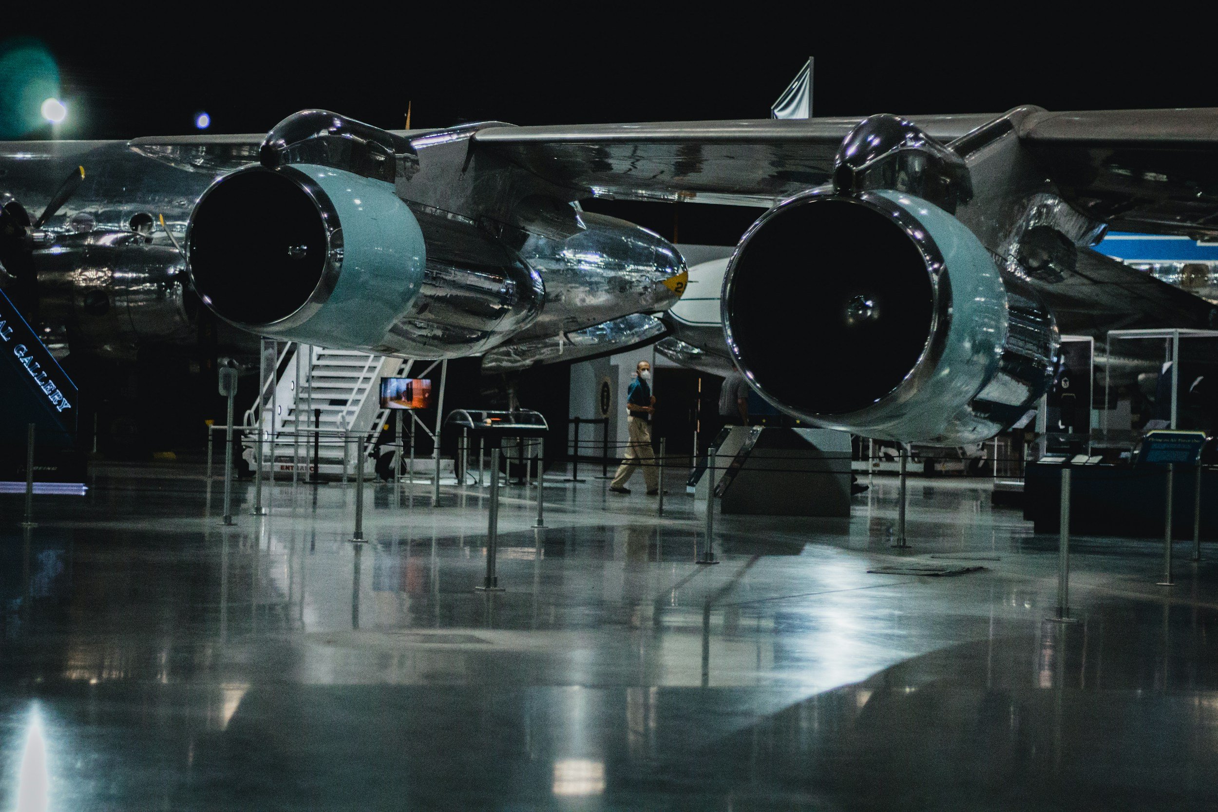 Photo of a shiny, large aircraft on display at an indoor exhibition at night, with a person wearing a mask and observing near the aircraft.