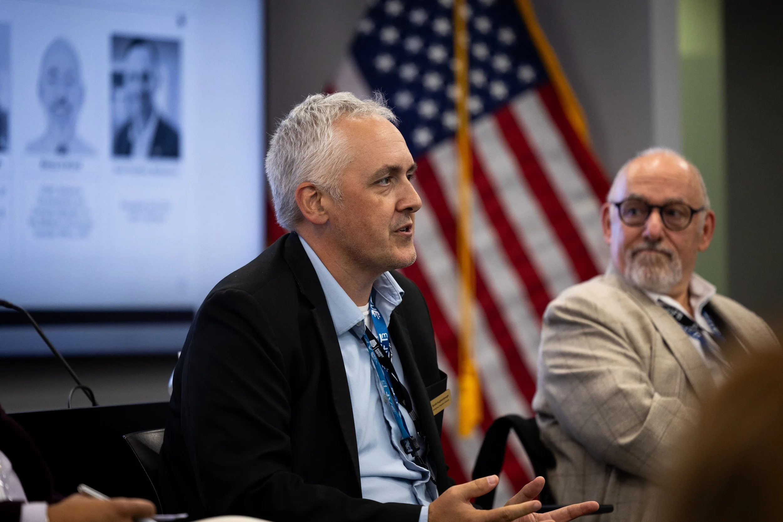 A man with gray hair and a dark suit is speaking at a conference or meeting, with an American flag in the background. Another man with glasses and a beard is seated next to him, listening. A presentation slide with headshots is visible on a screen to