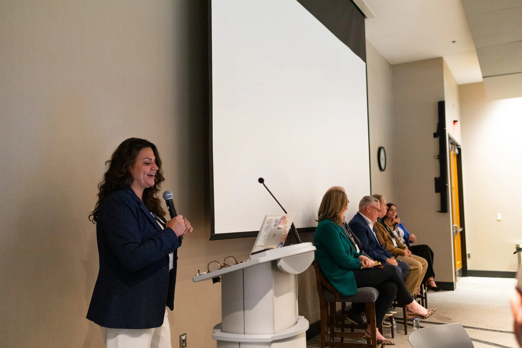 A woman with dark, curly hair is speaking into a microphone at a panel discussion or conference, standing at a white podium with papers on it. Several people are seated behind her along a row of chairs, facing forward, in a room with beige walls, a l