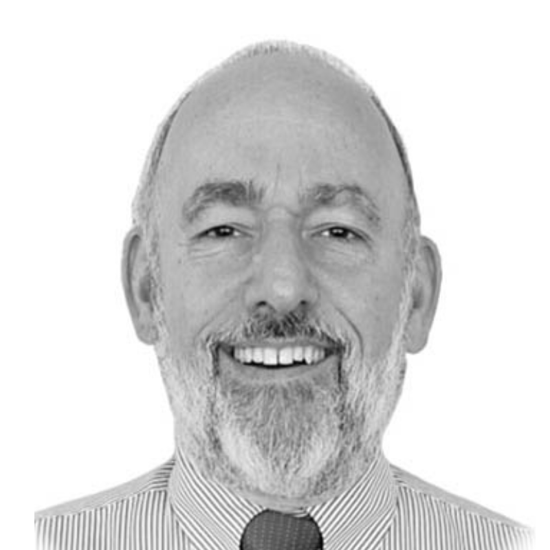 A black and white photo of a smiling man with short, light-colored hair, wearing a suit jacket over a collared shirt, standing in front of a background with diagonal wooden panels.