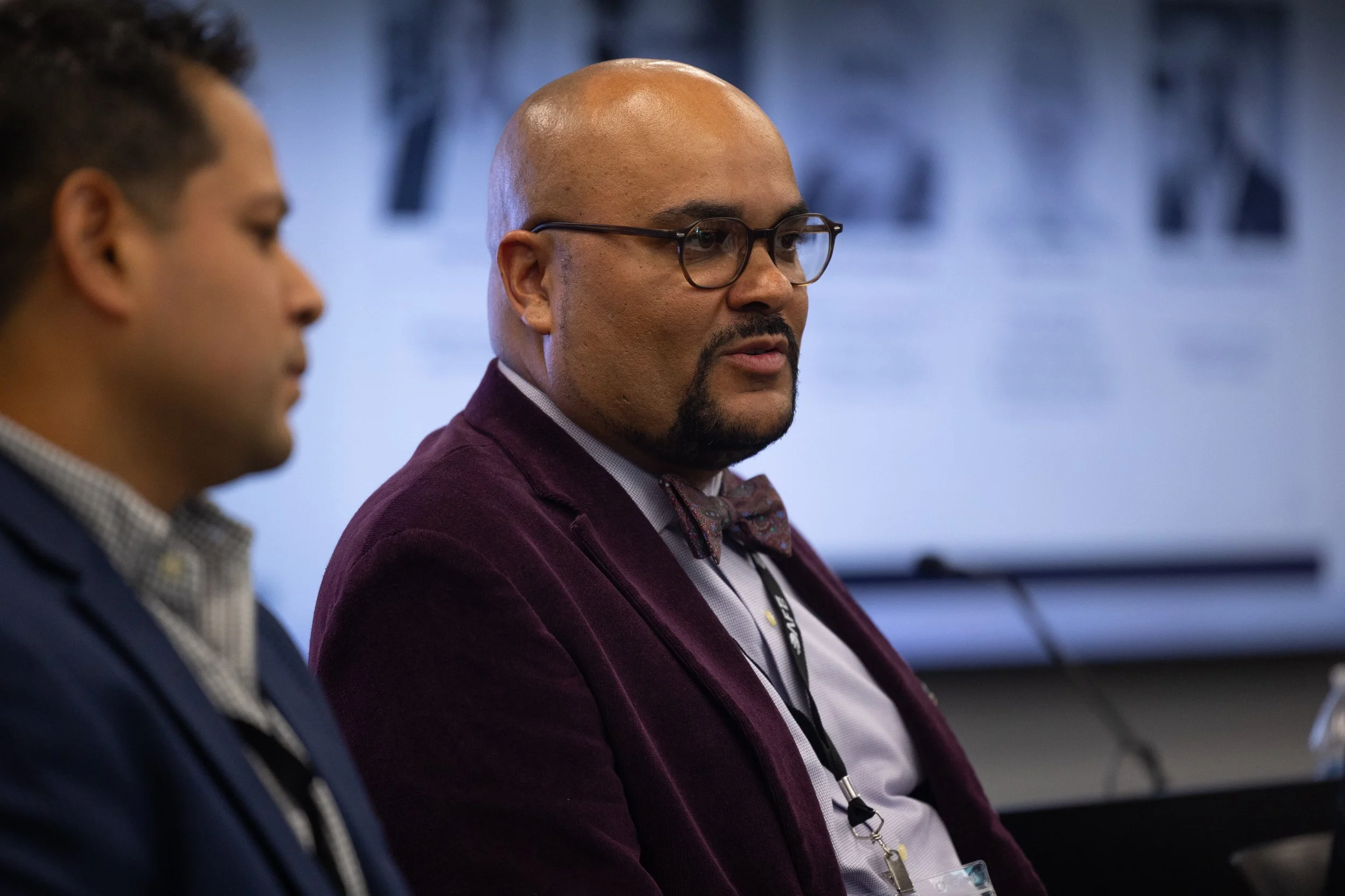 Two men sitting side by side, one with glasses and a bald head, wearing a maroon blazer, and the other with dark hair, in a setting that appears to be a conference or meeting room with a blue background.