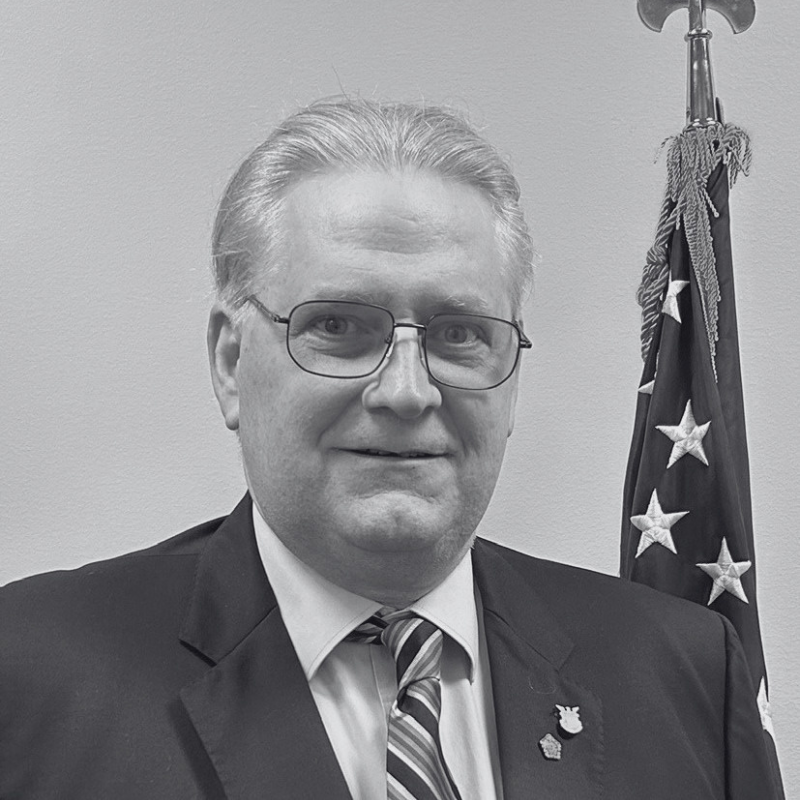 Portrait of a smiling man with short gray hair, wearing a checkered shirt and a dark jacket, standing outdoors near white pillars.