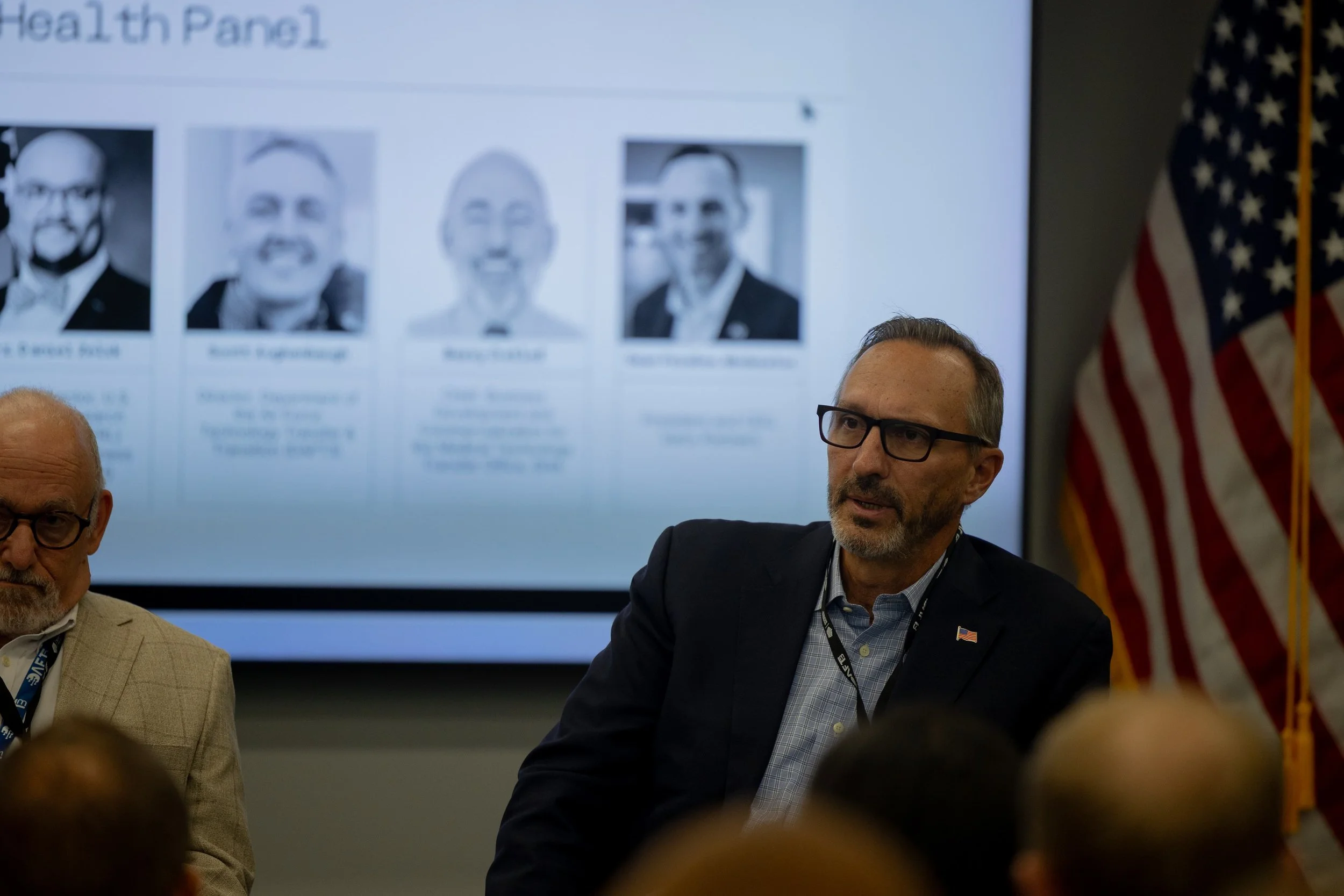 People participating in a discussion or meeting, with a presentation slide showing black and white photos of individuals in the background, and an American flag partially visible on the right side.