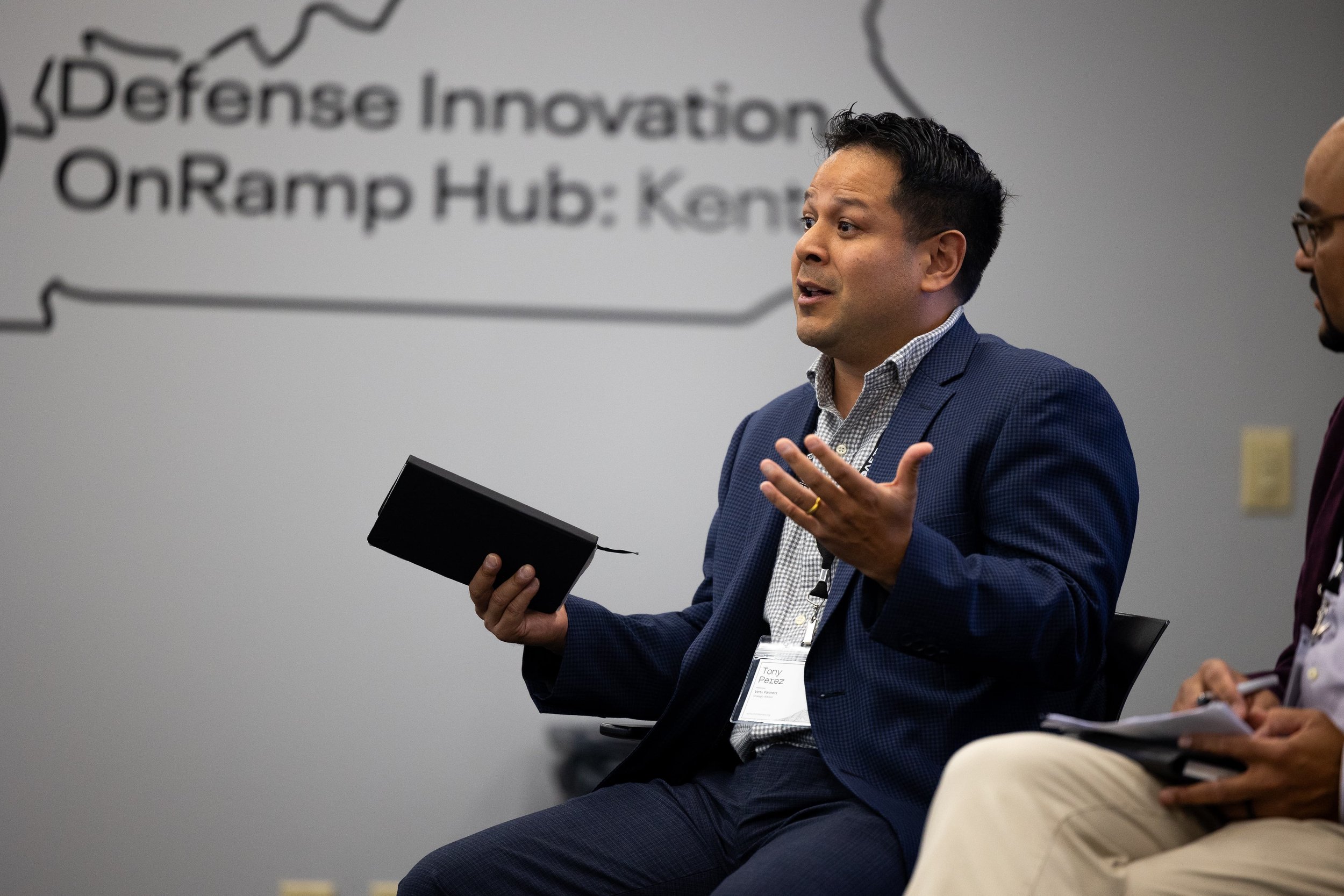 Man in a blue blazer giving a presentation or speech, holding a tablet, in a conference room with a blurred background showing a presentation slide.