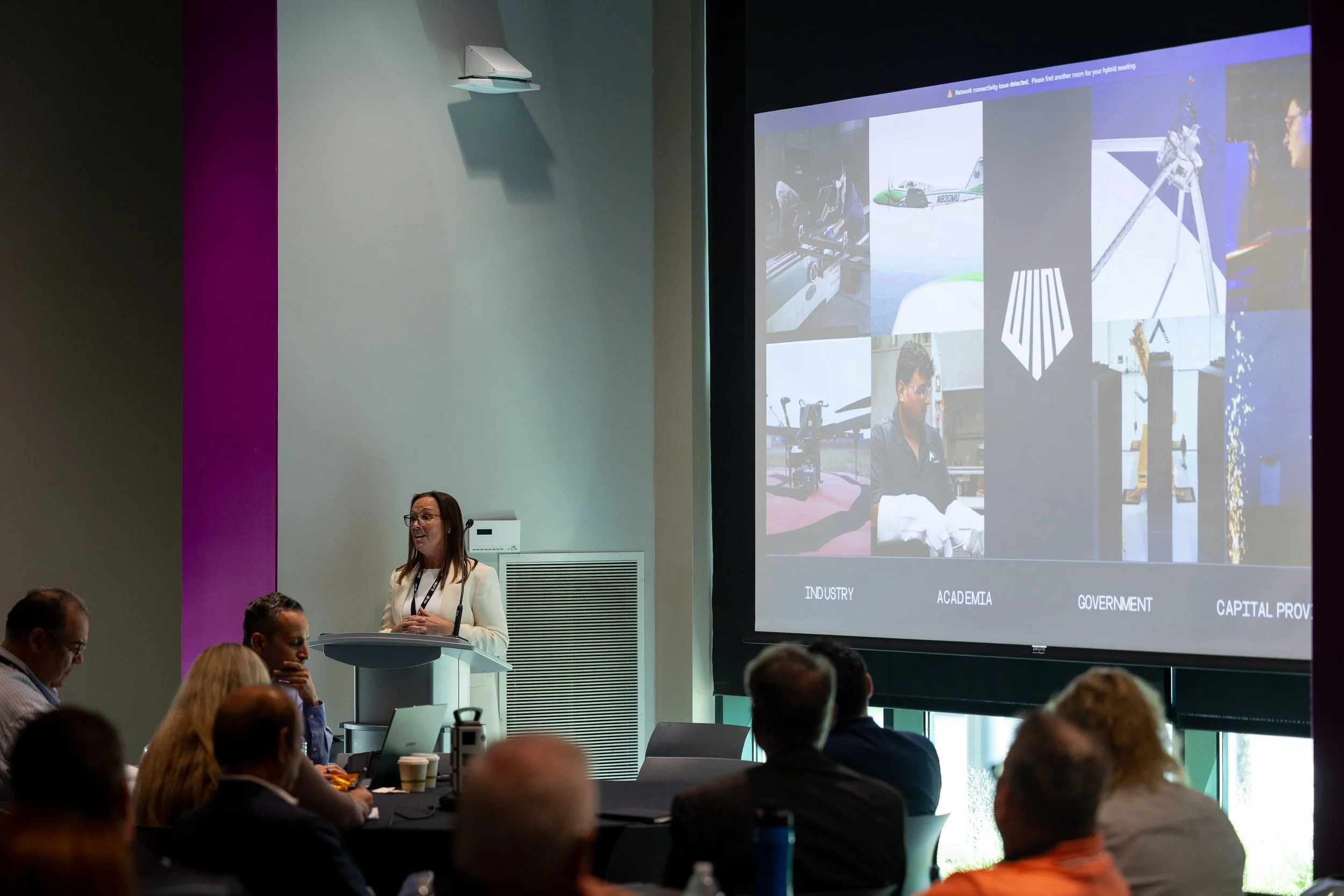 A woman standing at a podium giving a presentation in a conference room. A large screen behind her displays a slide with images and text, while audience members are sitting at tables listening.