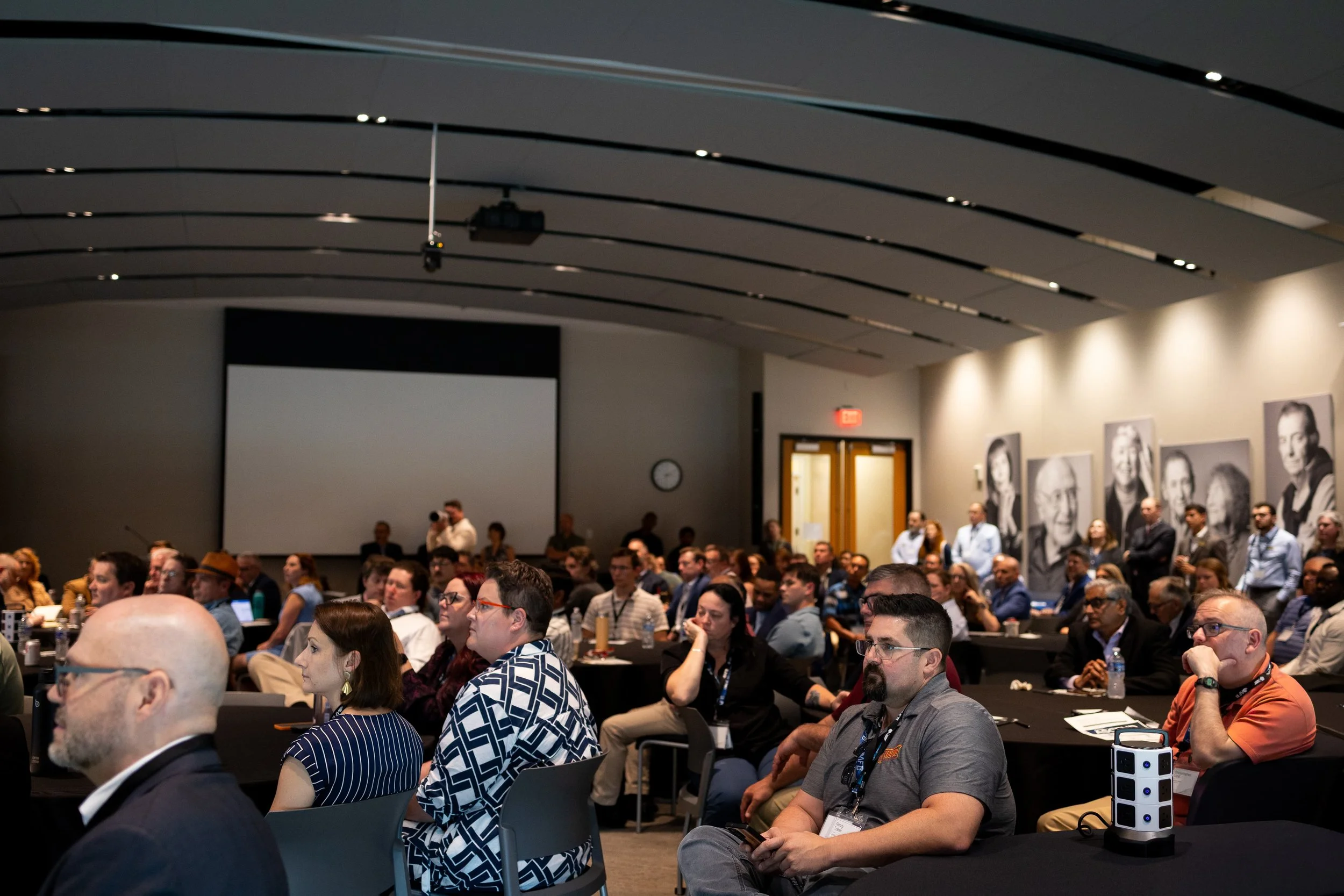 A large group of people attending a conference in a modern auditorium. The audience is seated at tables, listening attentively. The room has a high, curved ceiling with black paneling and small lights. Portrait photographs of various individuals are 