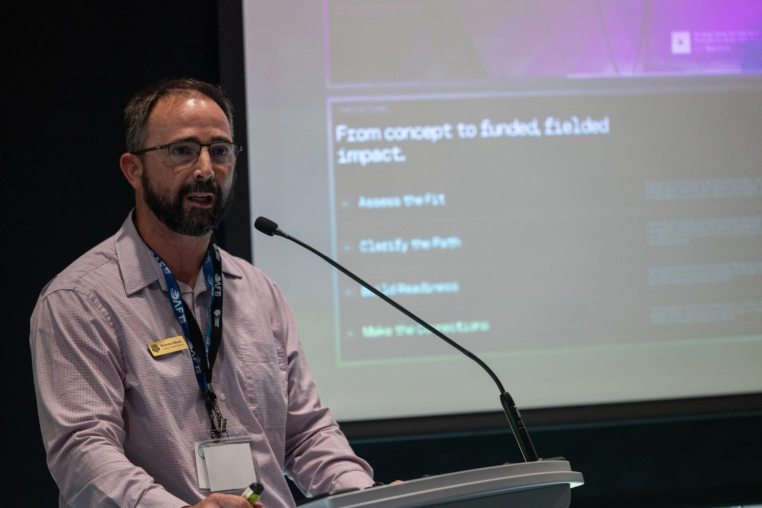 A man with glasses and a beard wearing a pink checkered shirt and a lanyard, standing at a podium and speaking into a microphone, with a presentation slide projected behind him.