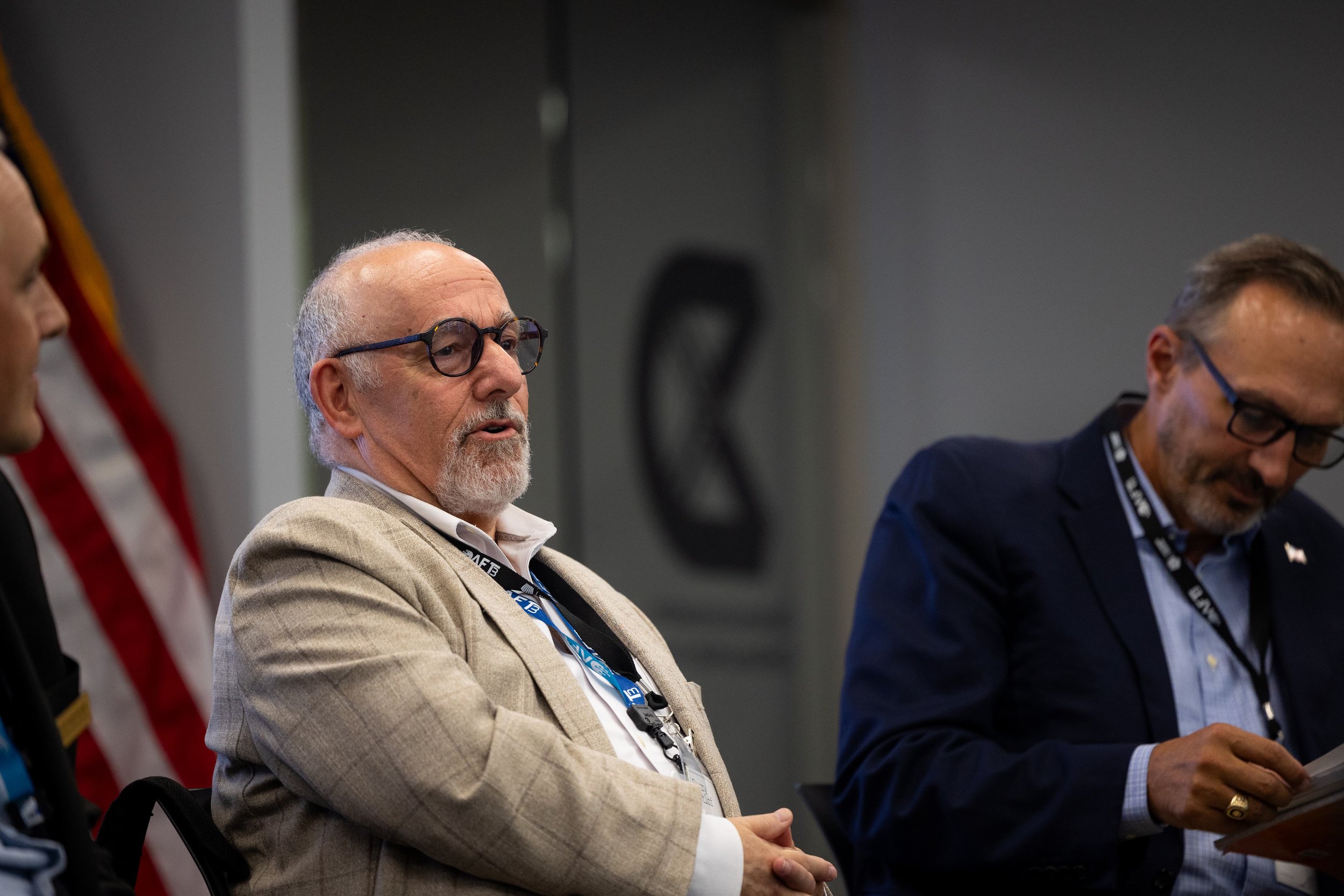 Three men seated at a panel, with the central figure being an older man with a white beard and glasses wearing a beige blazer. Two other men, one in a dark suit with glasses and another partially visible, are also present.