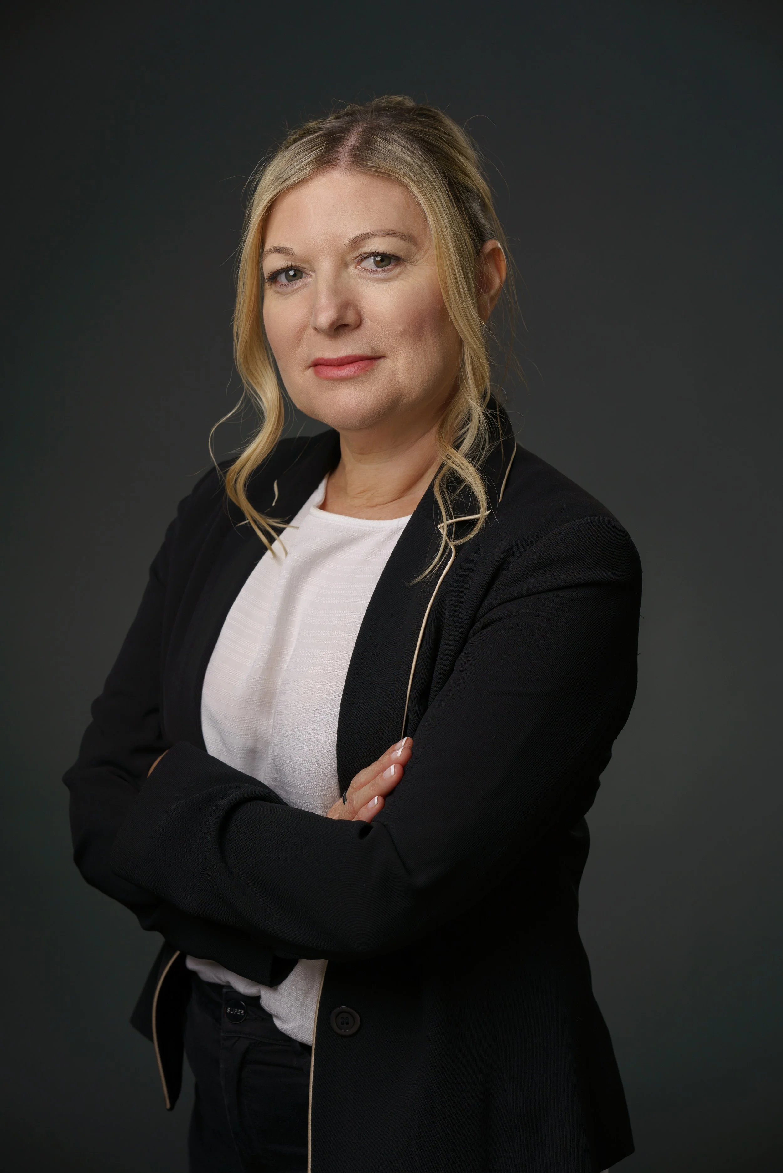A woman with blonde hair in a loose wave, dressed in a black blazer with gold piping and a white top, posing with her arms crossed against a dark background.