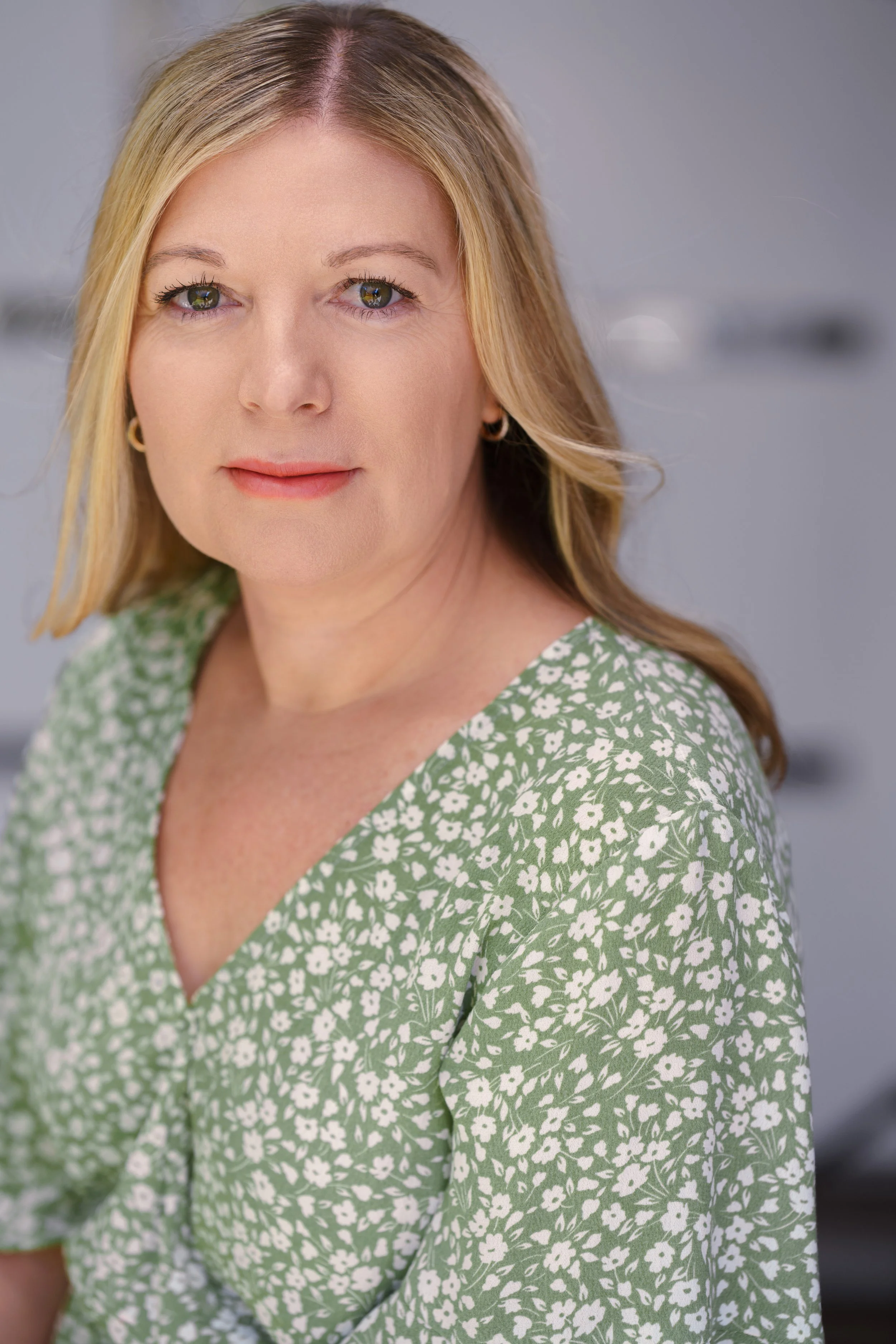 A woman with blonde hair, wearing a green blouse with white floral pattern, looking at the camera with a neutral expression, against a gray background.