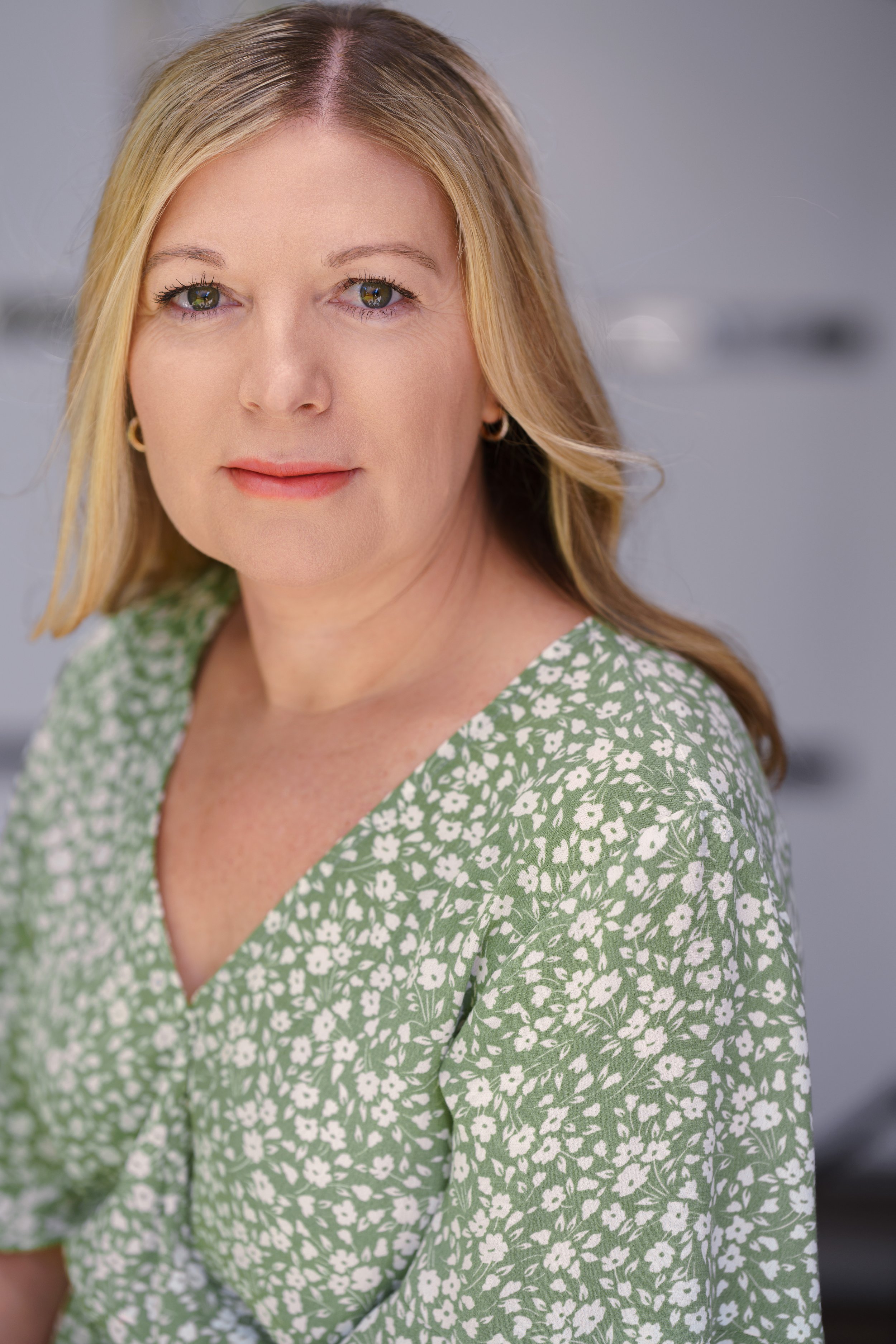 A woman with blonde hair wearing a green dress with a white floral pattern, looking into the camera.