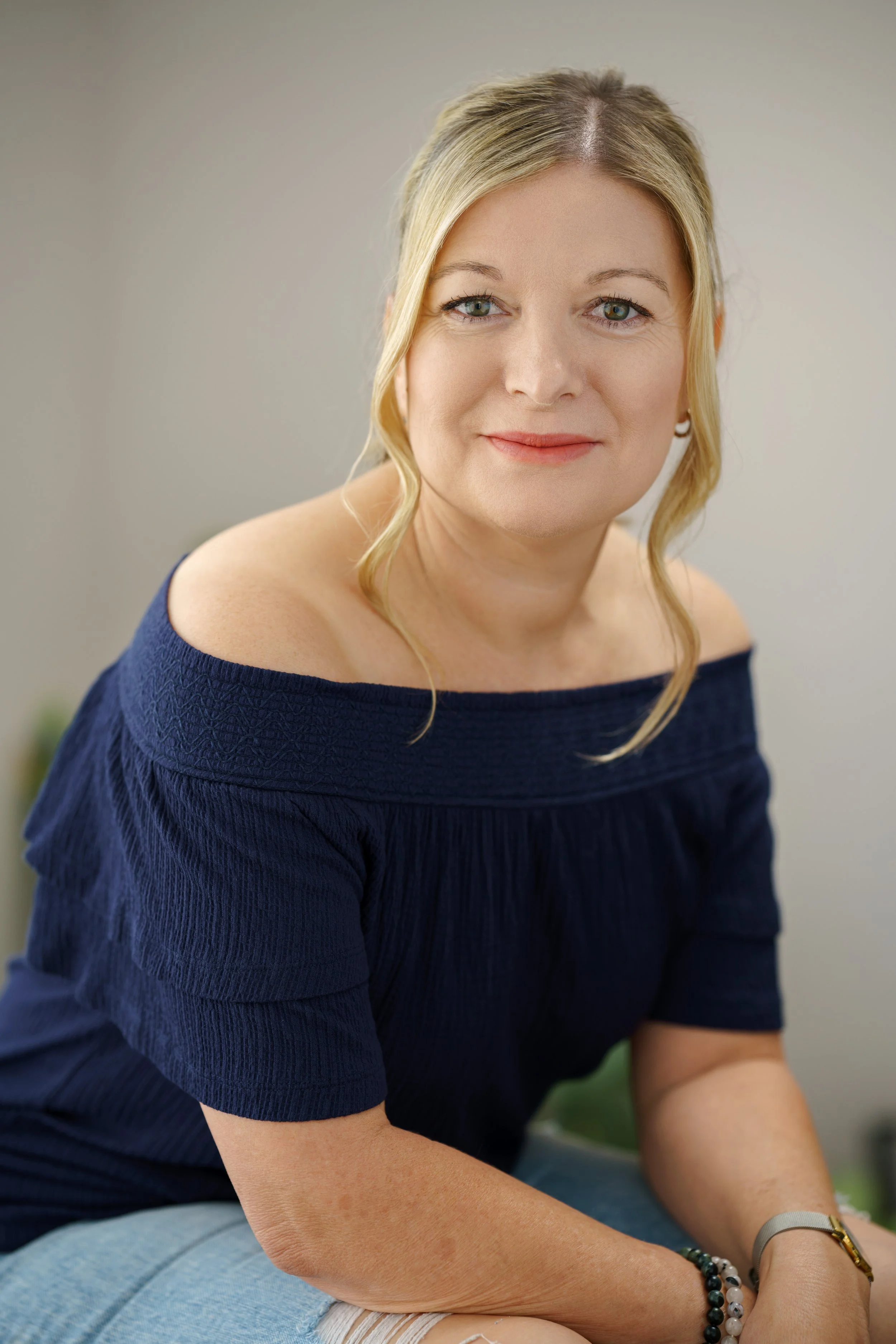 A woman with blonde hair and light skin, wearing a navy blue off-shoulder top, smiling and looking at the camera. She is sitting with her arms resting on her lap, wearing bracelets and a watch.