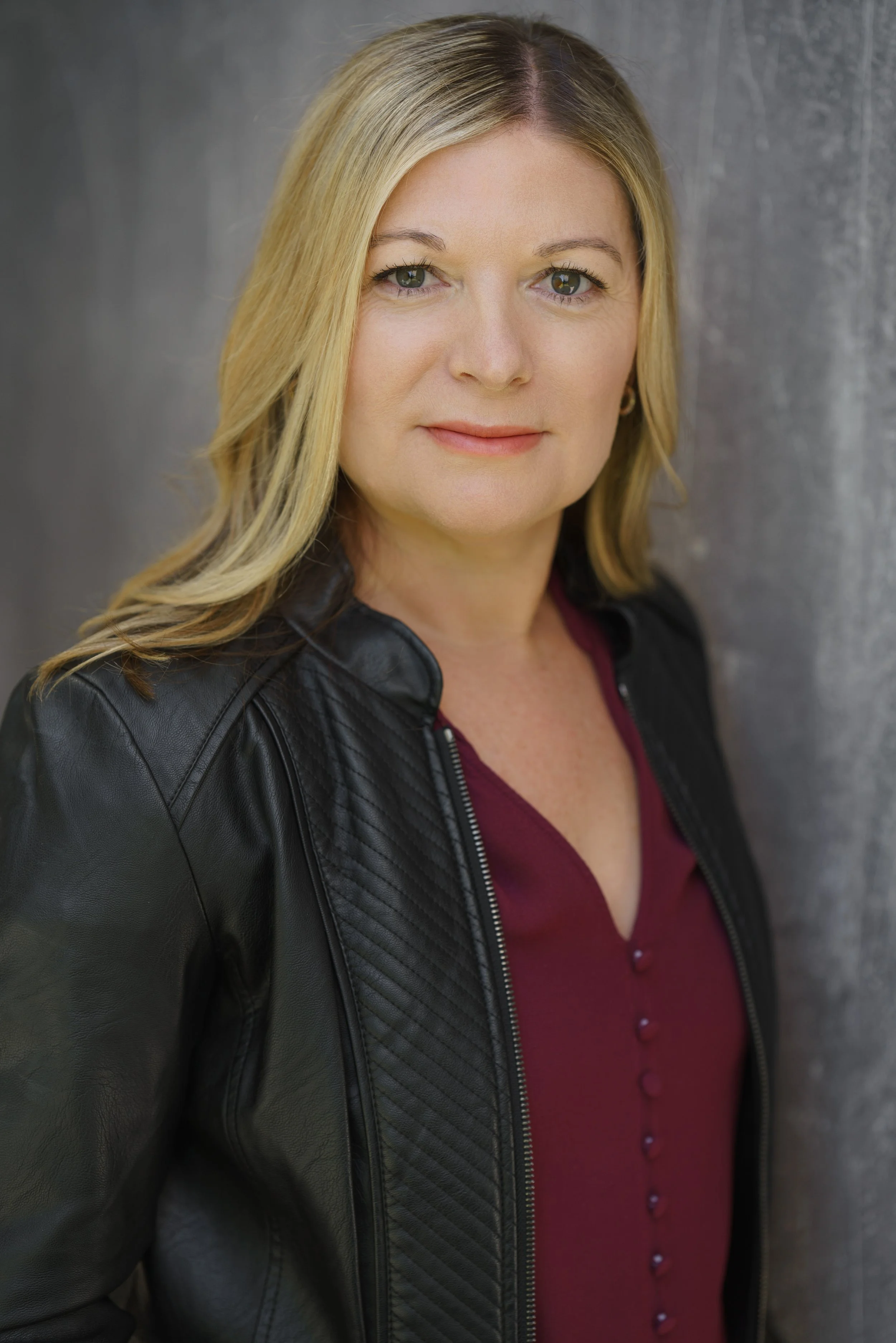 Portrait of a woman with blonde hair wearing a black leather jacket and a burgundy top, standing against a gray textured background.
