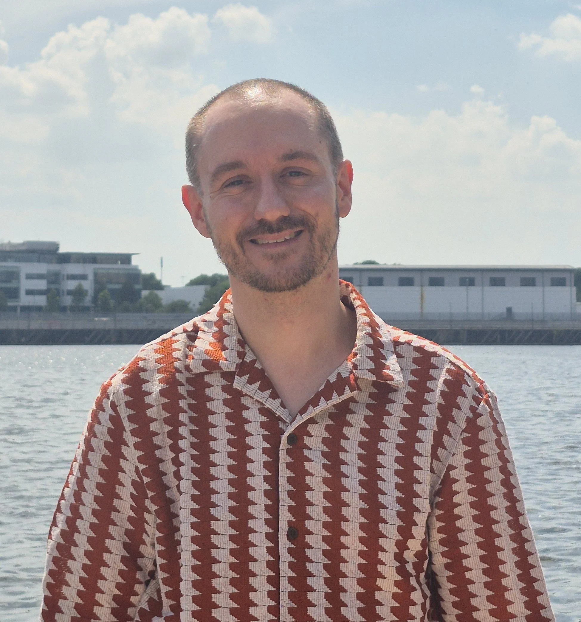 A smiling man with short hair and a beard, wearing a patterned button-up shirt, standing outdoors near water with modern buildings and a cloudy sky in the background.