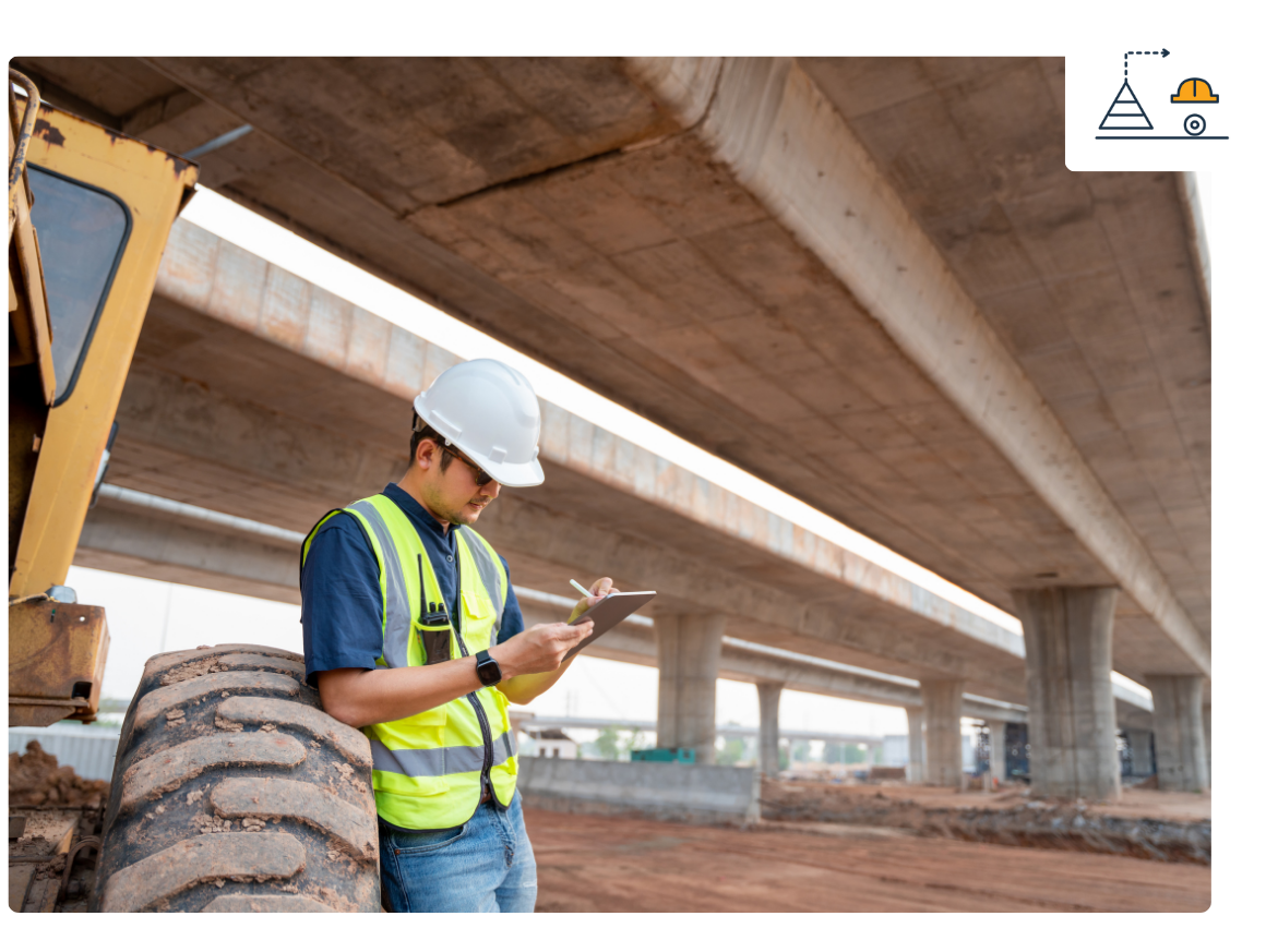 Construction worker with a white hard hat and safety vest inspecting under a bridge with a tablet in hand.