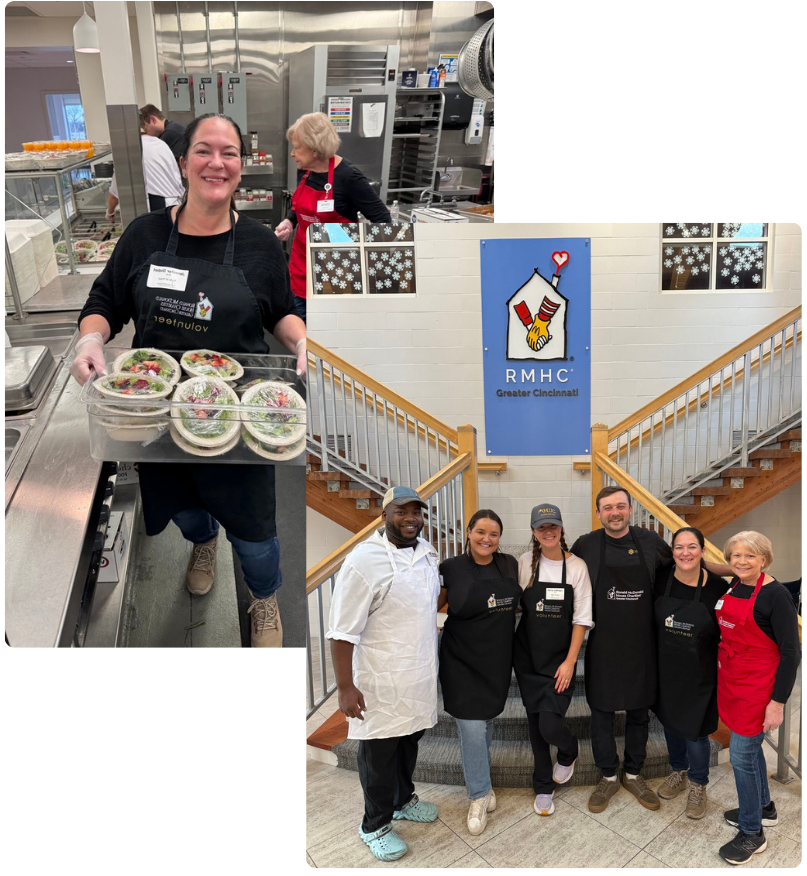 A woman in a black apron holding containers of salad in a kitchen, with two women in red aprons cooking in the background and a group of five people standing on a staircase in front of a sign that reads 'RMHC Greater Cincinnati.'