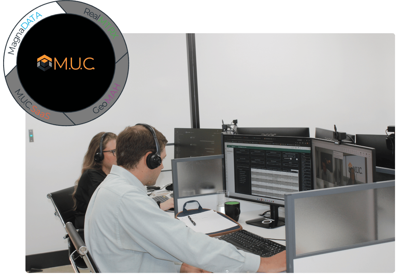 Two people working at desks with multiple computer monitors in an office, with a M.U.C logo and circular diagram on the wall.