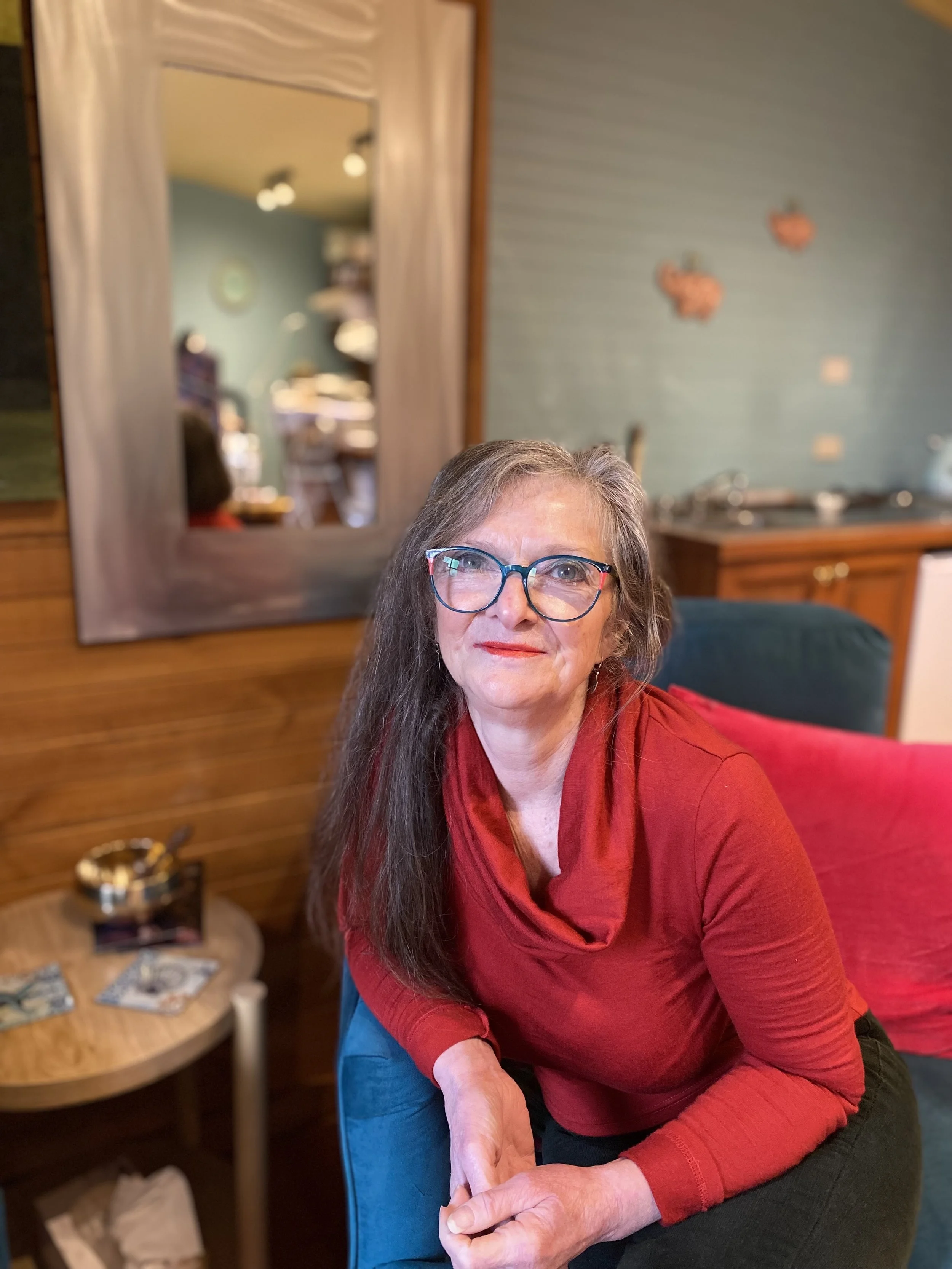 An elderly woman with glasses and a red sweater sitting in a living room, smiling, with a mirror reflecting a room with a clock and books in the background.