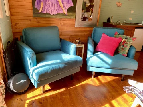 Living room with two teal armchairs, one with a red pillow and the other with a floral pillow, wooden floor, and a wooden wall with artwork. There's a small side table and a robot vacuum nearby.