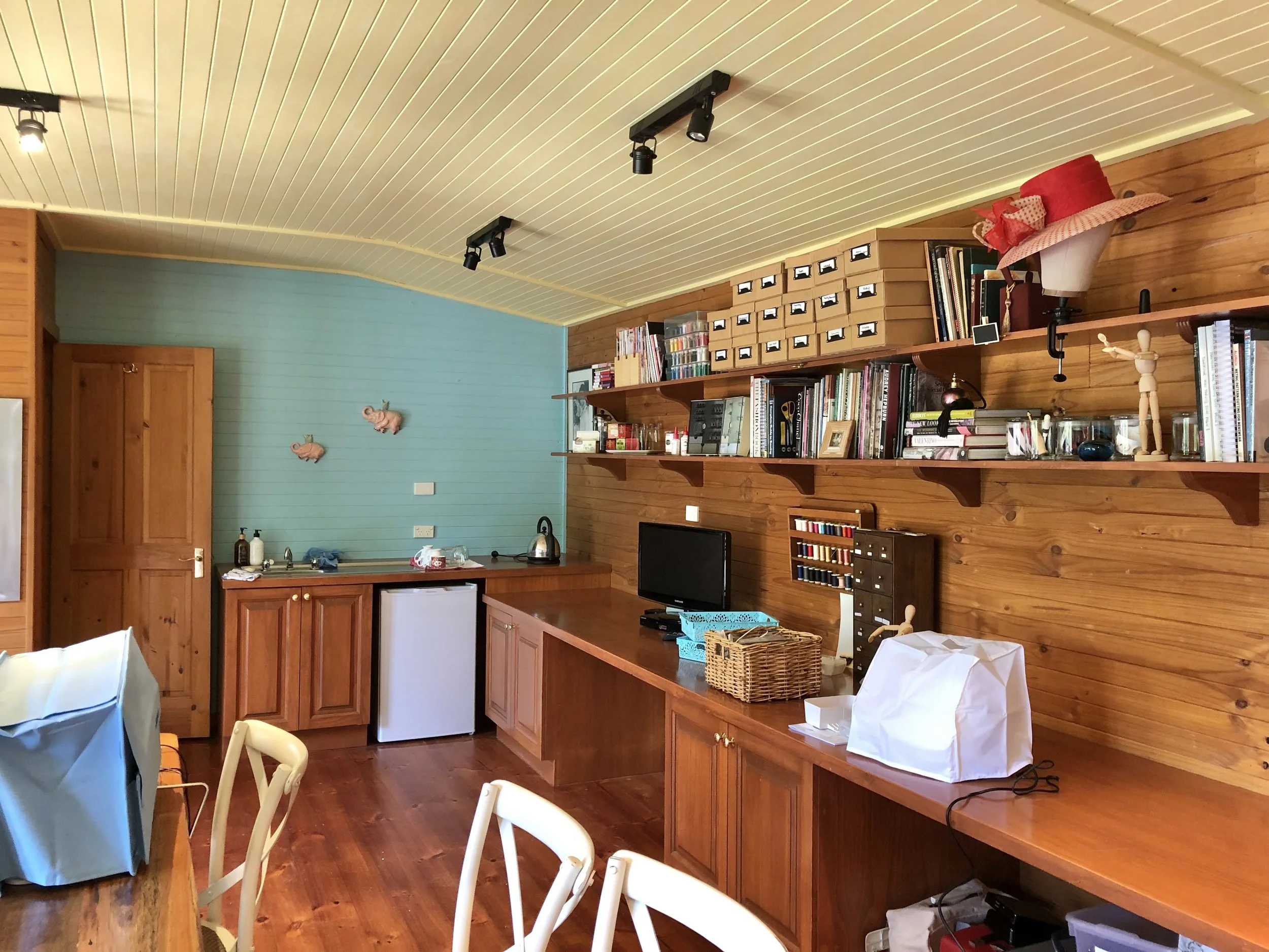 A room with wood-paneled walls and a cream-colored, beadboard ceiling. There are bookshelves filled with books and storage boxes, and various decorative items including a pink floral hat and small pig ornaments on the wall. The room has a small kitchenette area with a sink, a mini refrigerator, and a kettle. There are several chairs around a table, and various paper bags and items on the countertops.