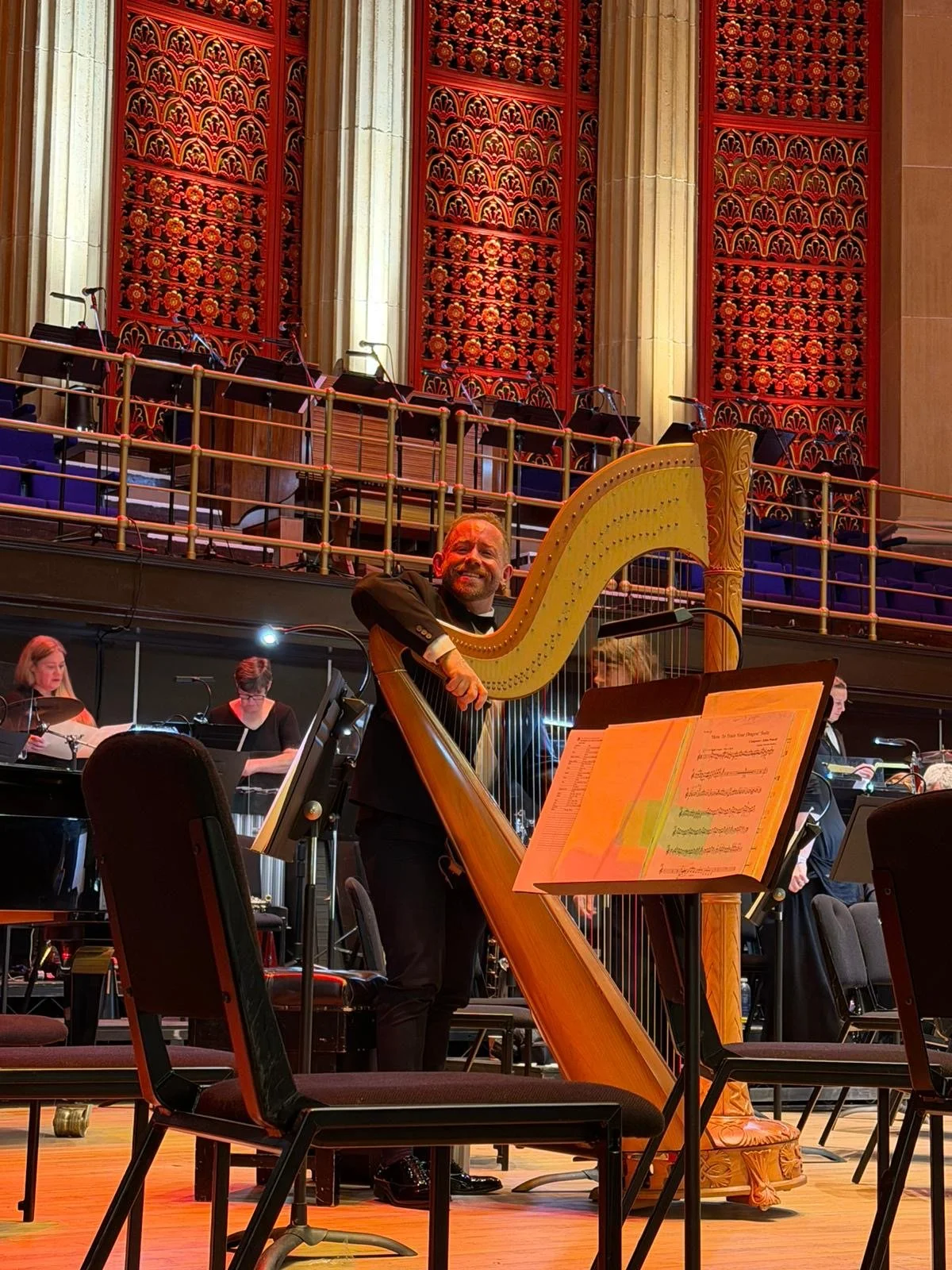 A man in a tuxedo smiling and playing a harp during a classical concert in a concert hall with ornate red wall decorations and other musicians in the background.