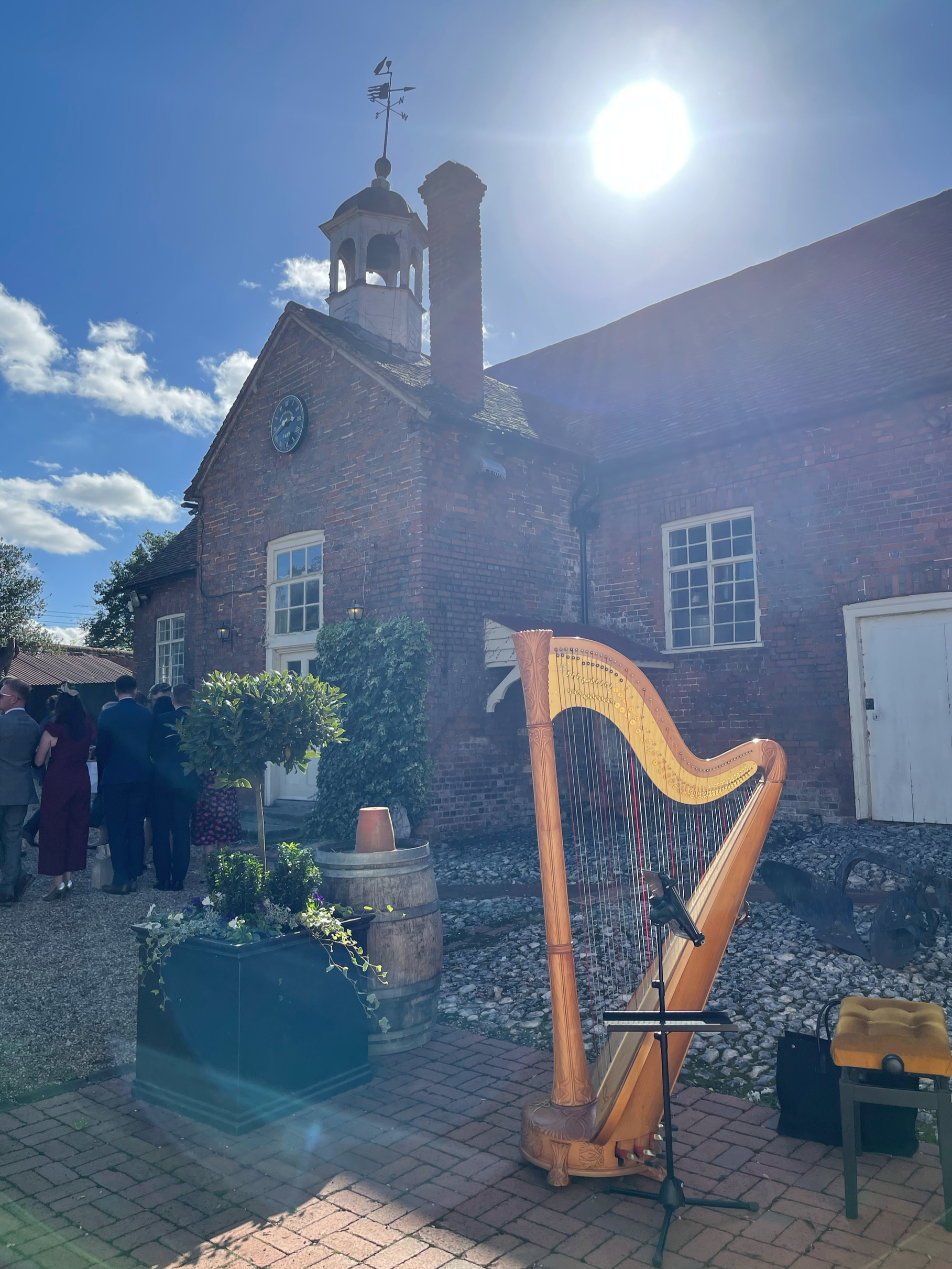 A harp, a microphone stand, and a padded stool are set up outside a brick building with a clock and a small tower with a weather vane on top, under a bright sun in a mostly clear sky. People are gathered nearby.
