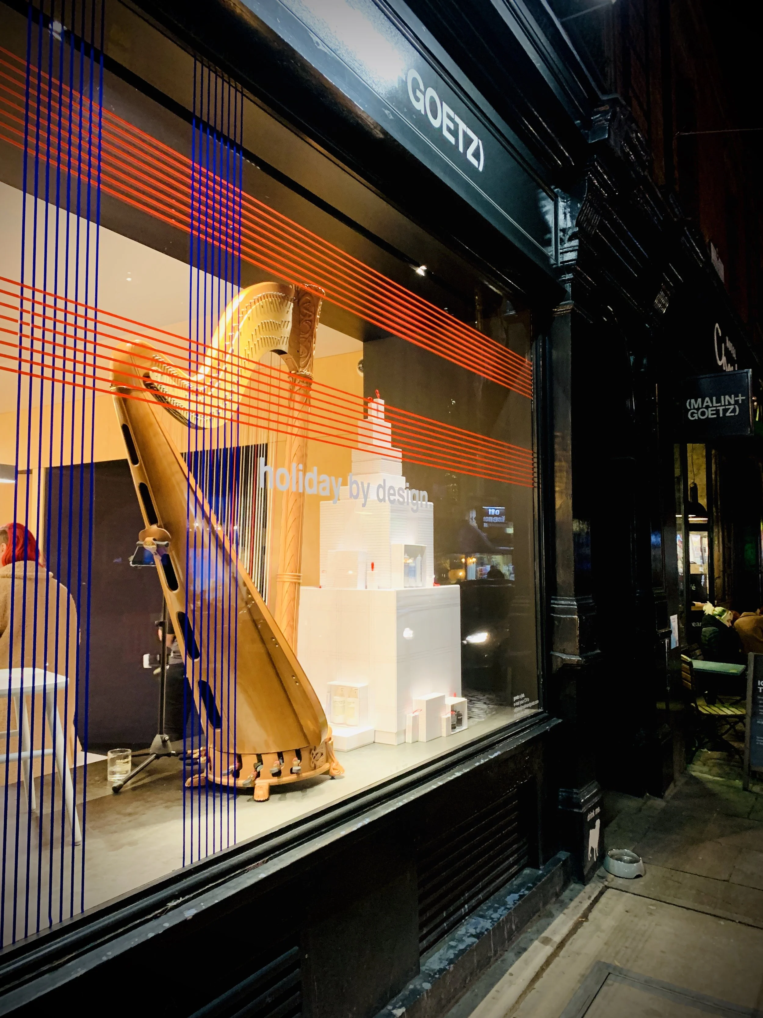 Store window display with a wooden harp and abstract decor, with orange and blue lines across the glass, and signs that read 'GOETZ' and 'MALIN + GOETZ' at night.