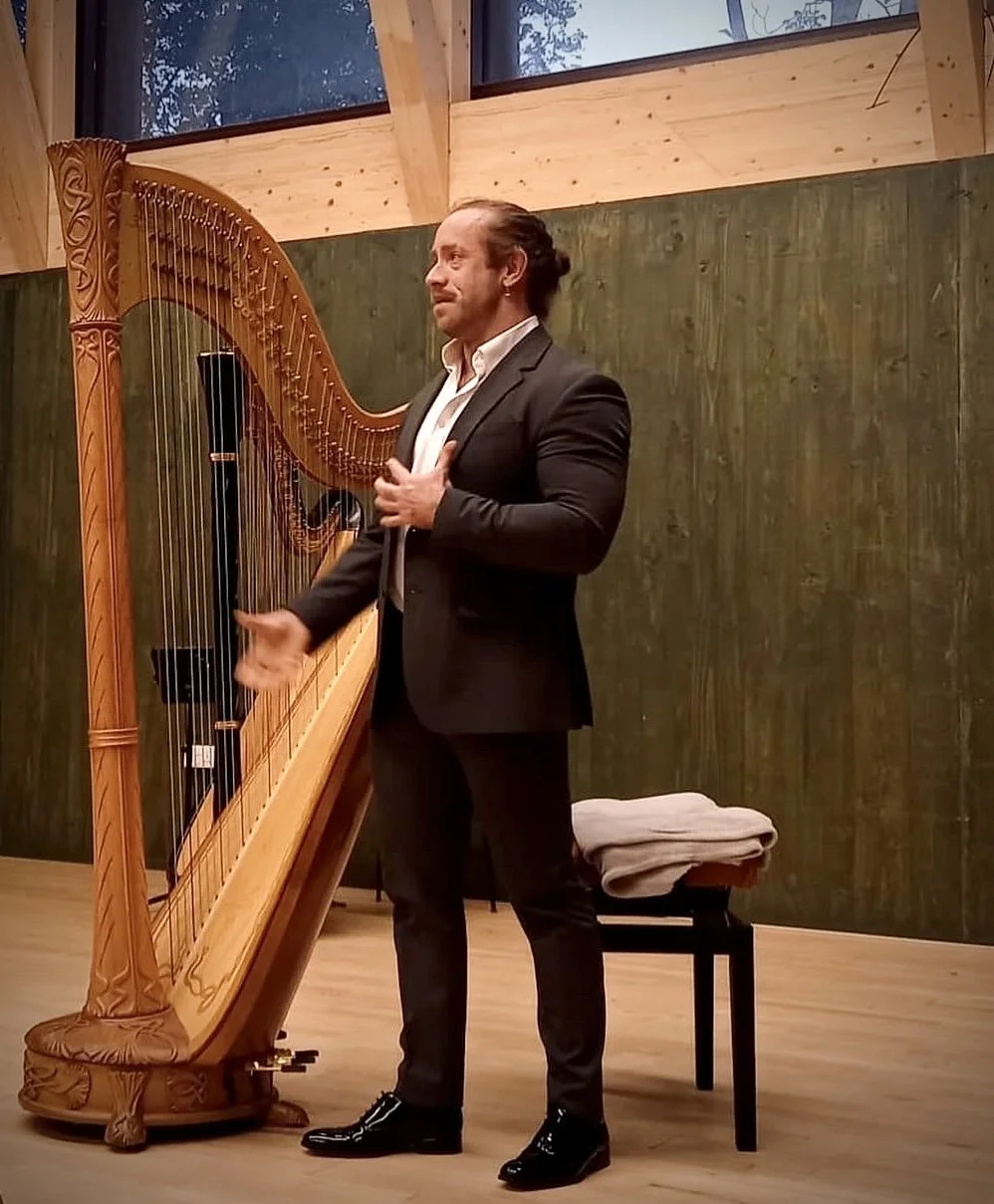 A man in formal attire playing a harp in a wooden room with large windows.