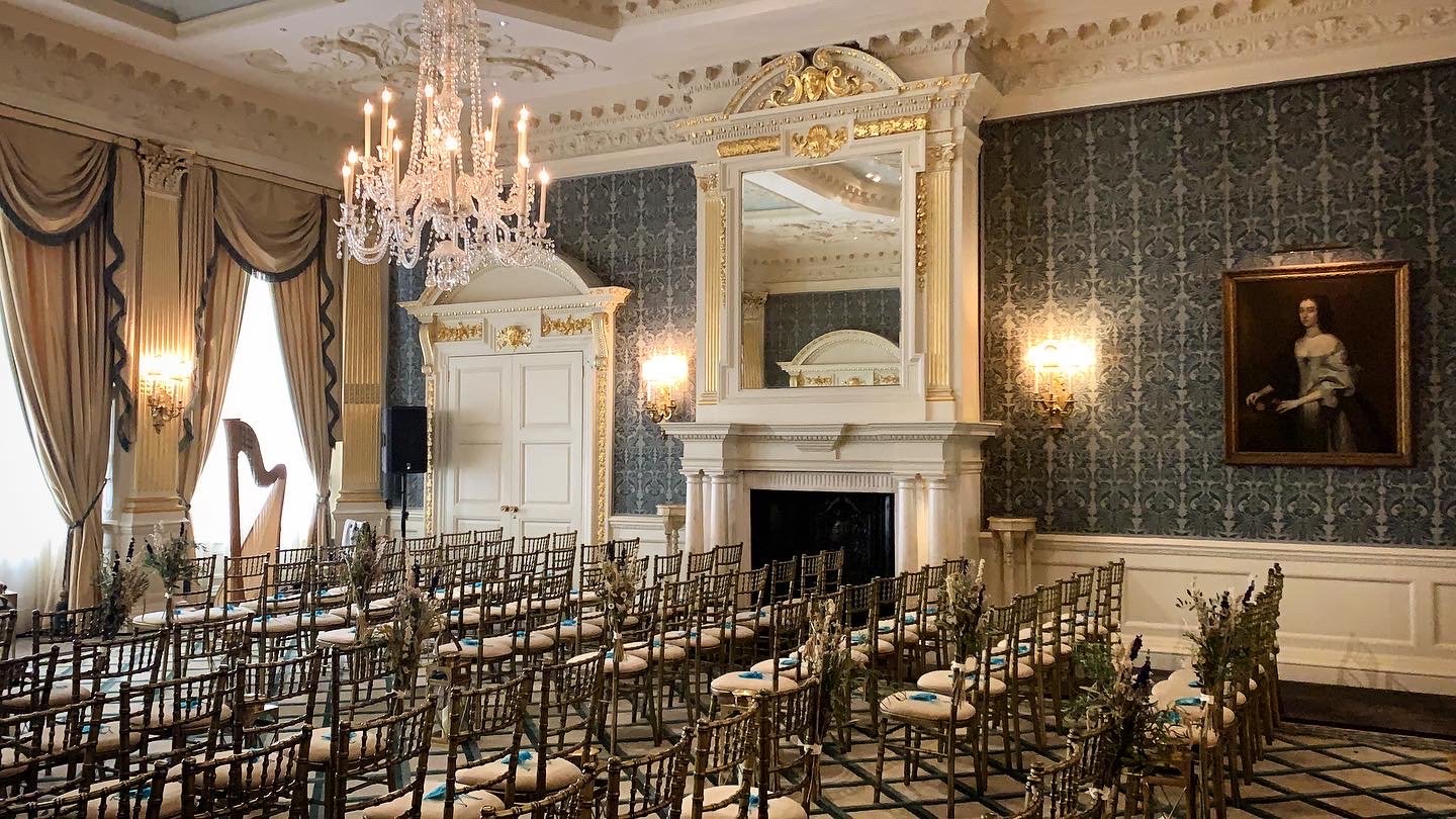 Elegant wedding or event setup in a luxurious room with ornate decor, crystal chandelier, rows of white chairs with floral arrangements, fireplace with large mirror, and dark patterned wallpaper.