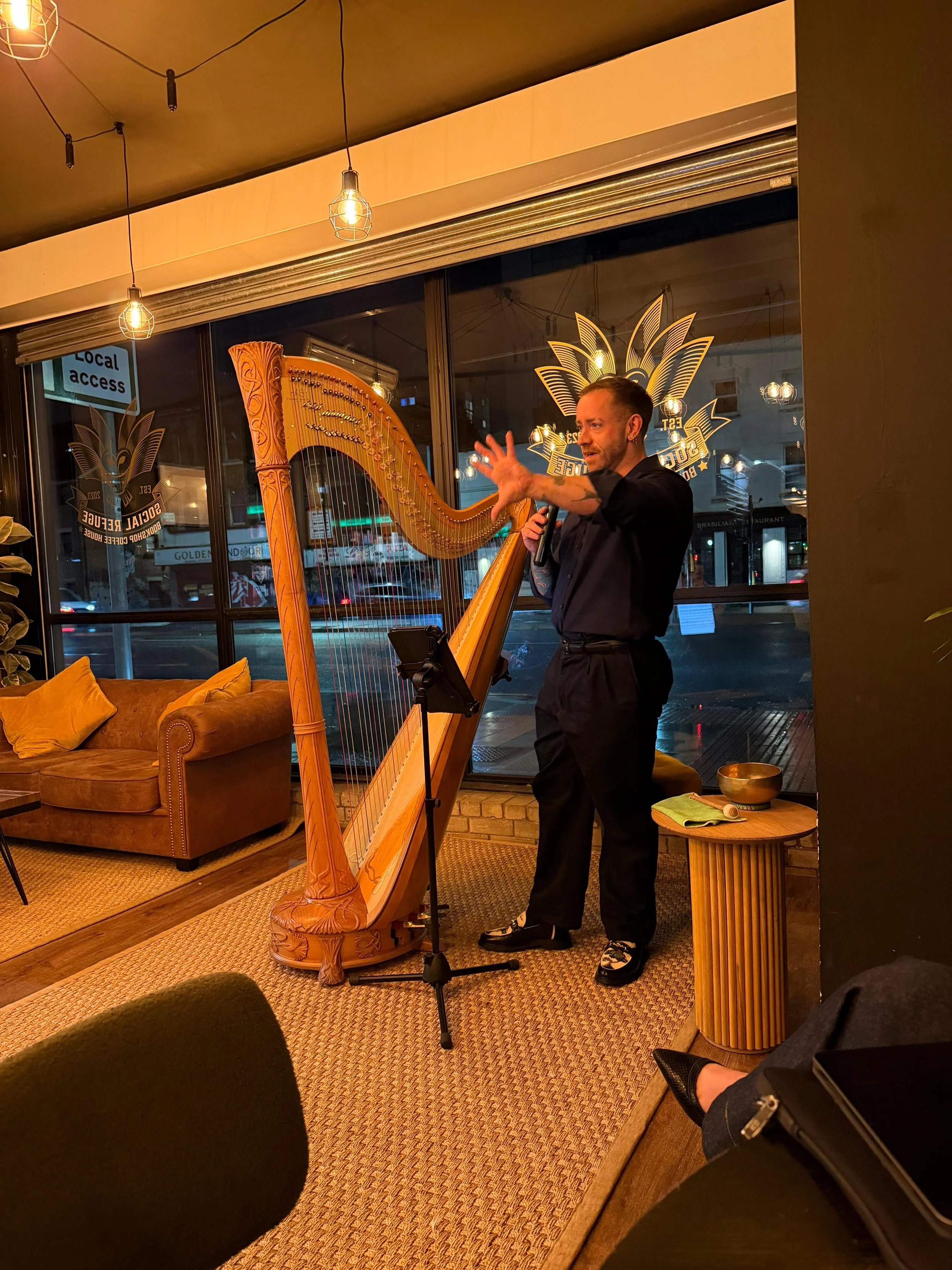 Man playing a harp inside a cozy, warmly-lit cafe or restaurant at night, with orange cushions on a sofa and glass windows showing city street outside.