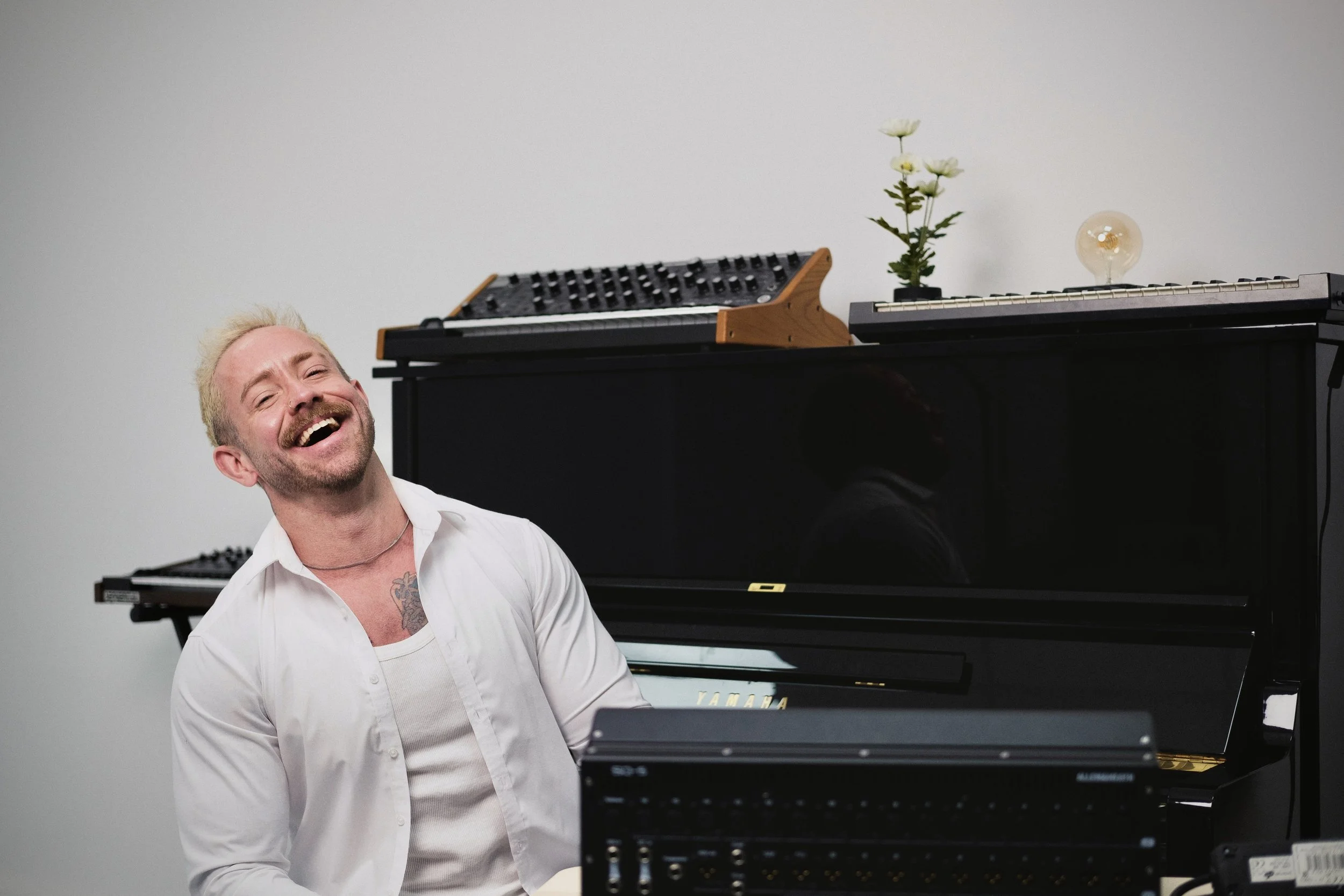 A man with blonde hair and a mustache, wearing a white shirt, is laughing and sitting in front of a black Yamaha piano. The piano has a vintage light bulb and a small plant with white flowers on top. The background is a plain, white wall.